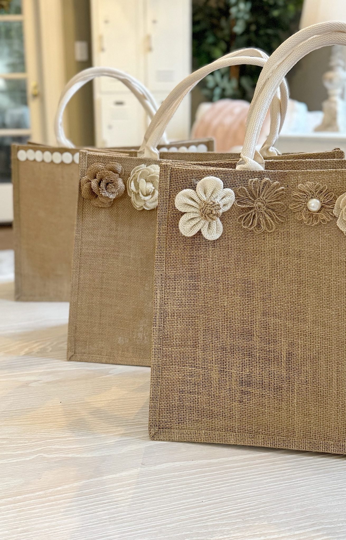 Three burlap tote bags with white handles are decorated with fabric flowers along the top edge, arranged in a row on a light wooden floor.