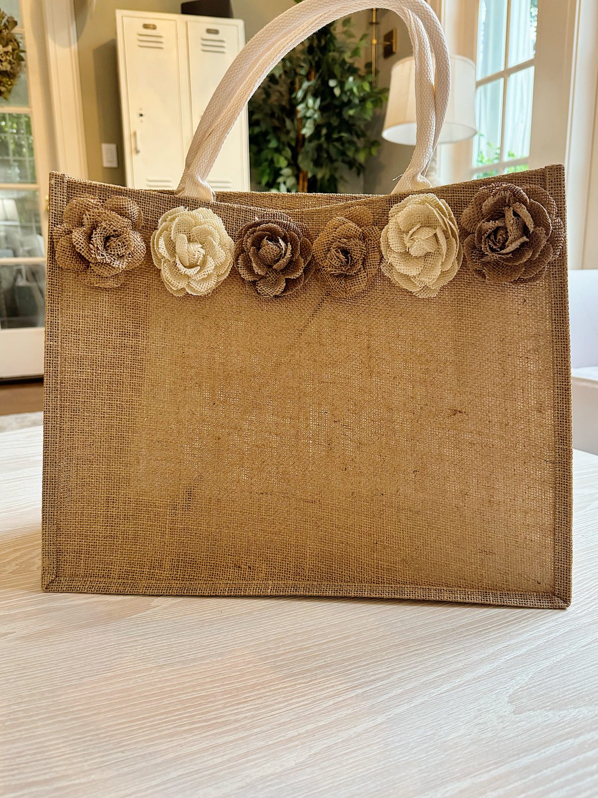 A beige jute tote bag with white handles, decorated with five fabric flowers in various neutral shades, sits on a light-colored wooden table indoors.