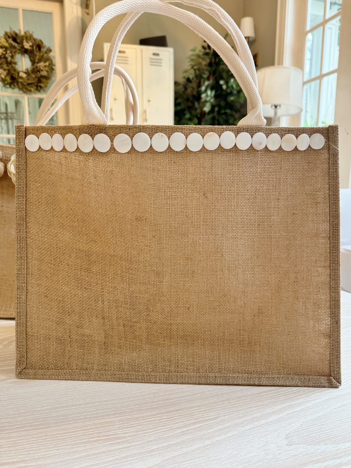 A burlap tote bag with white rope handles and a row of round white buttons along the top edge, placed on a light-colored surface indoors.