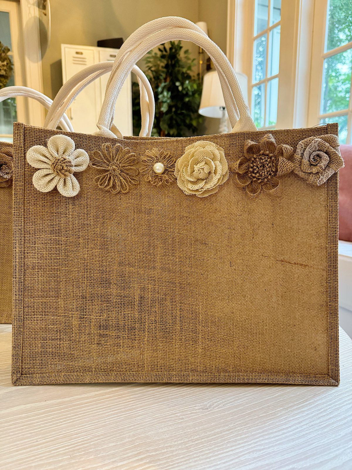 A jute tote bag with cream and brown fabric flowers attached along the top edge, displayed on a table indoors near a window.