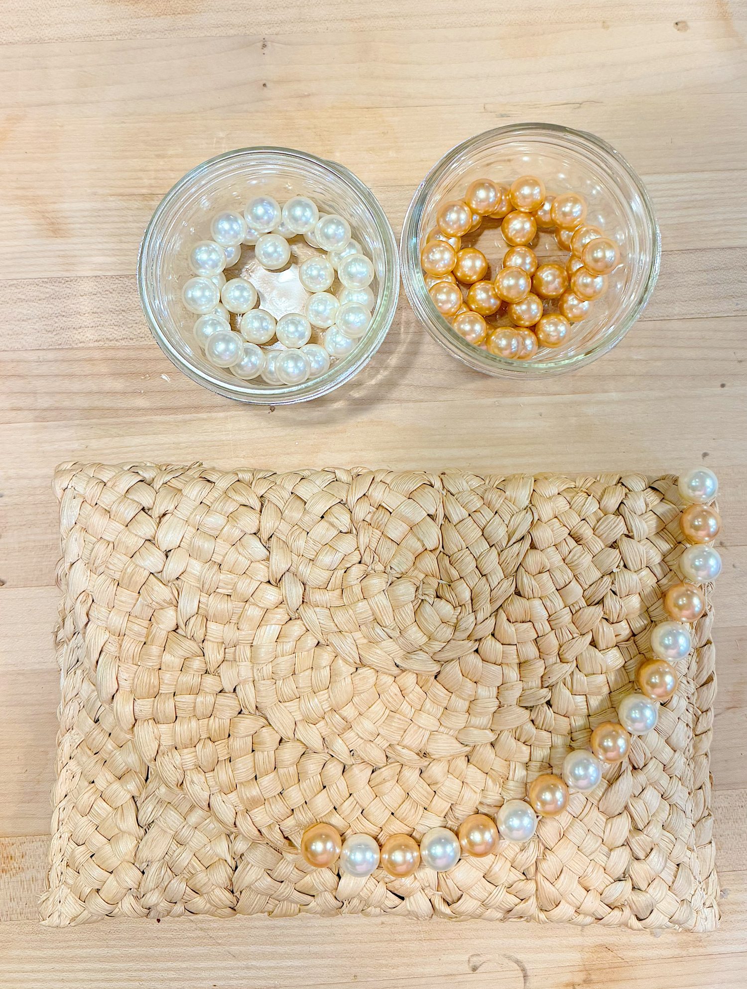 Two small glass bowls with white and gold pearls sit on a light wooden surface next to a woven clutch and a string of mixed pearls.