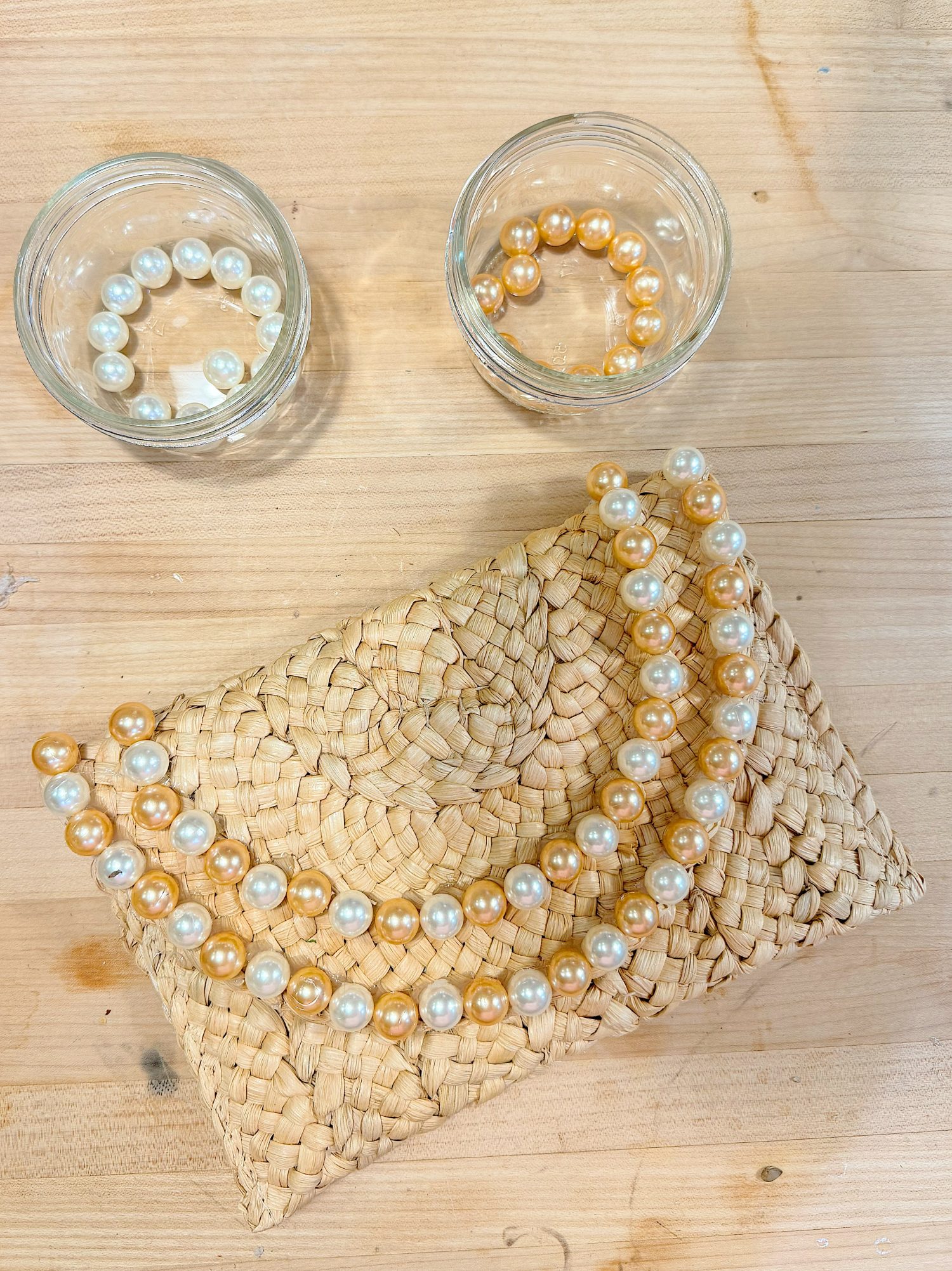 Two glass jars with pearl bead bracelets and a woven clutch with two pearl bead necklaces are arranged on a light wooden surface.