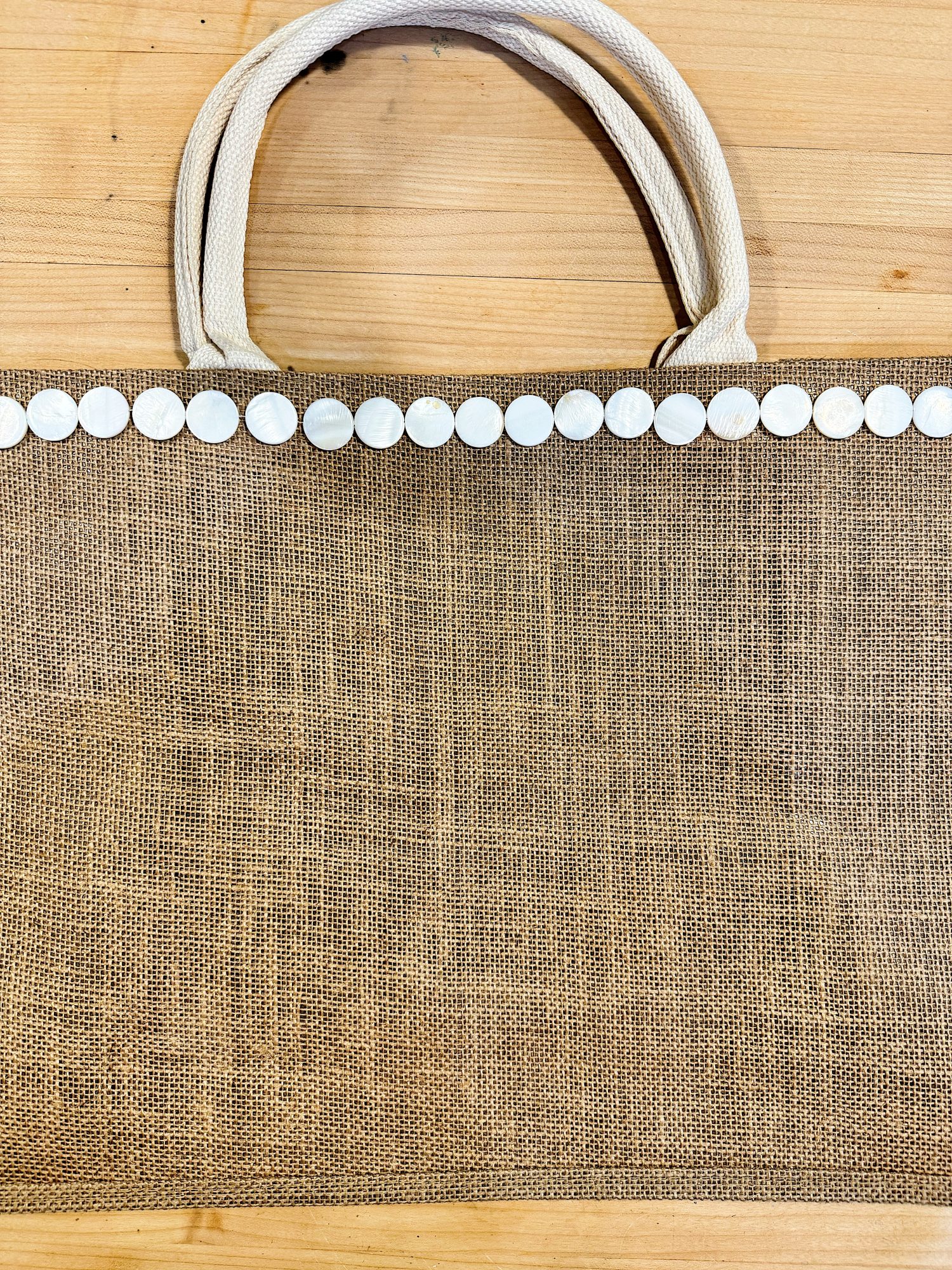 A jute tote bag with white circular decorations along the top edge and two white fabric handles, placed on a wooden surface.