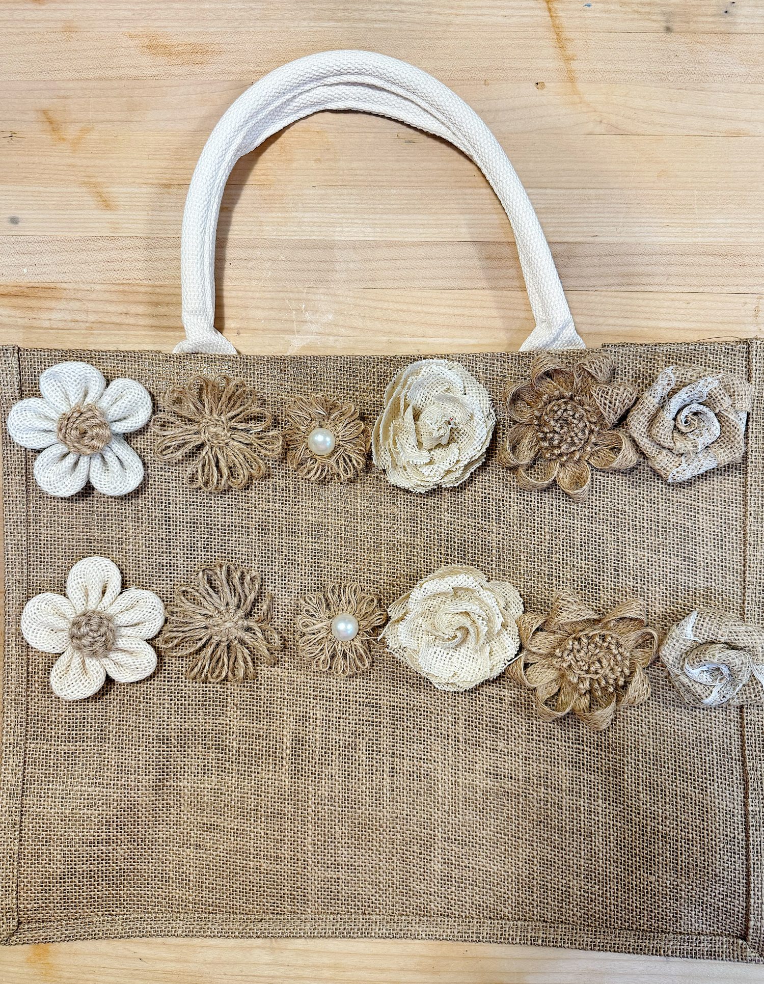 A burlap handbag with white rope handles, decorated with fabric and jute flowers, some featuring pearls, on a wooden surface.