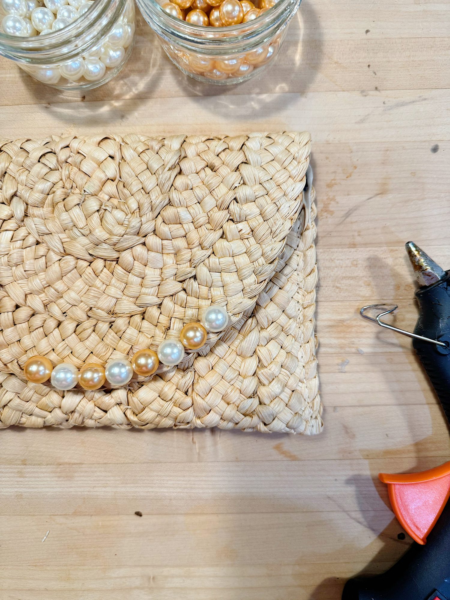 A woven straw clutch decorated with gold and white pearls sits on a wooden surface next to jars of beads and a hot glue gun.