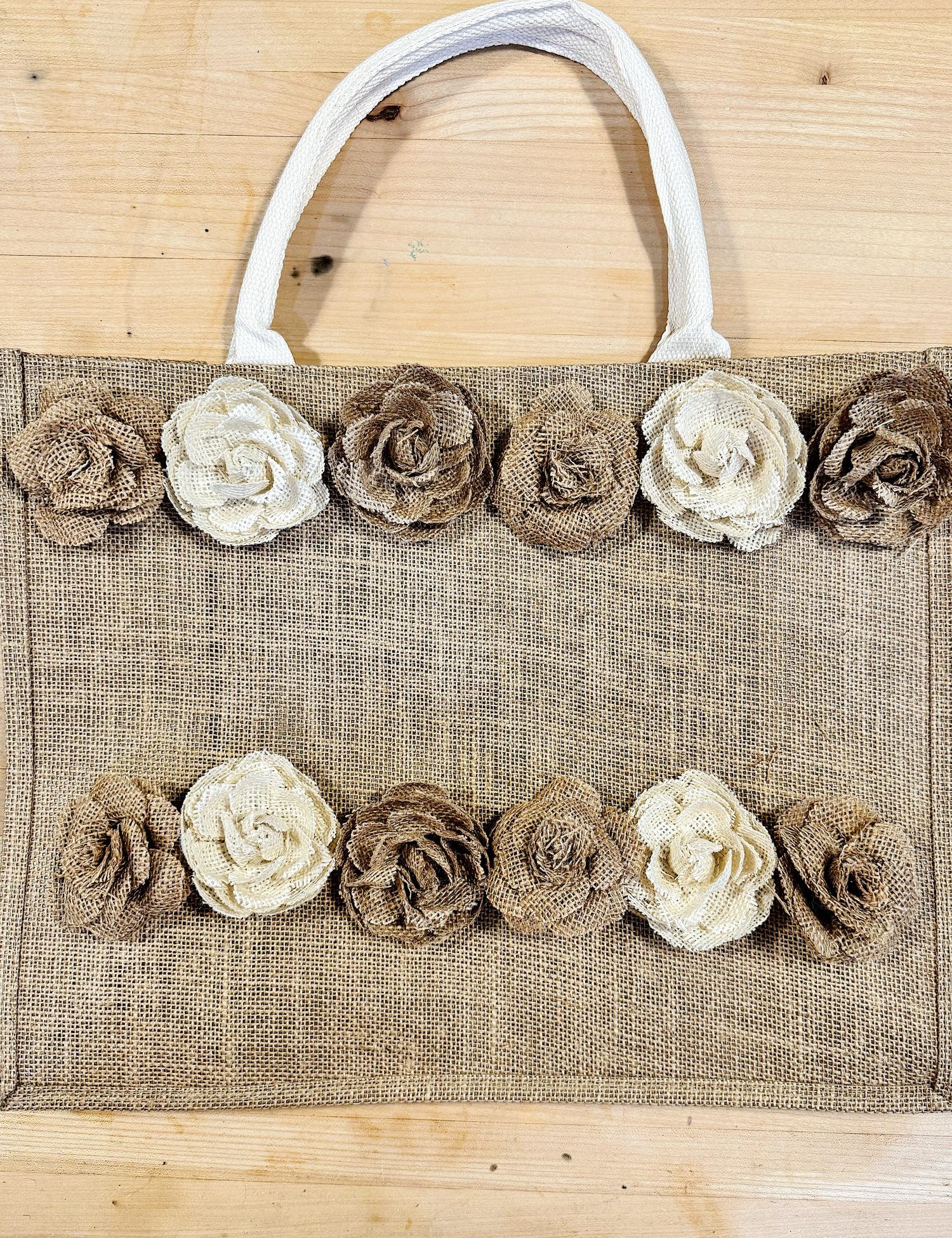 A burlap tote bag with two rows of decorative fabric roses in cream and brown shades, placed on a wooden surface.