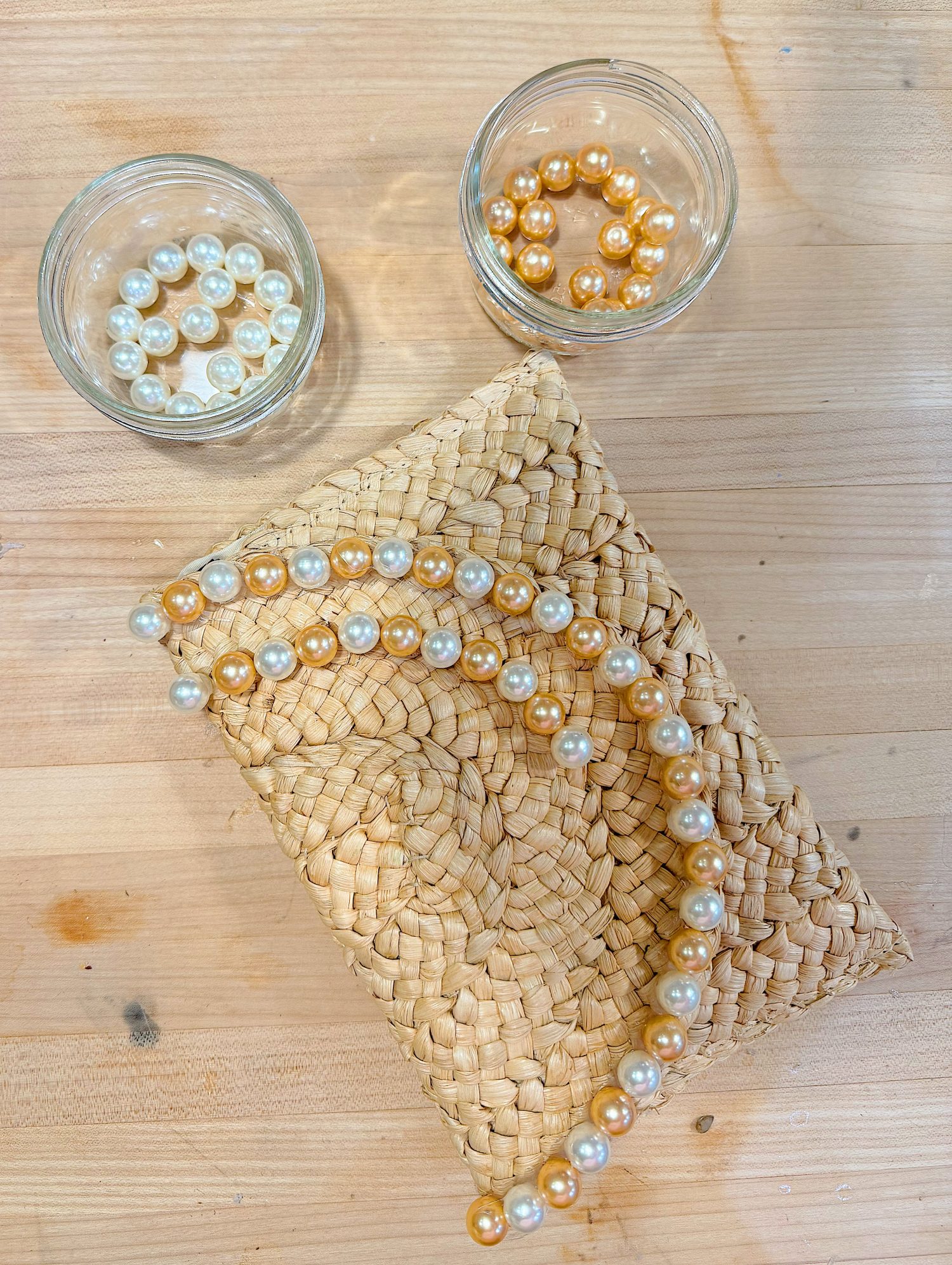Two glass jars with white and gold beads, and a braided straw pouch with a matching white and gold beaded necklace on a wooden surface.