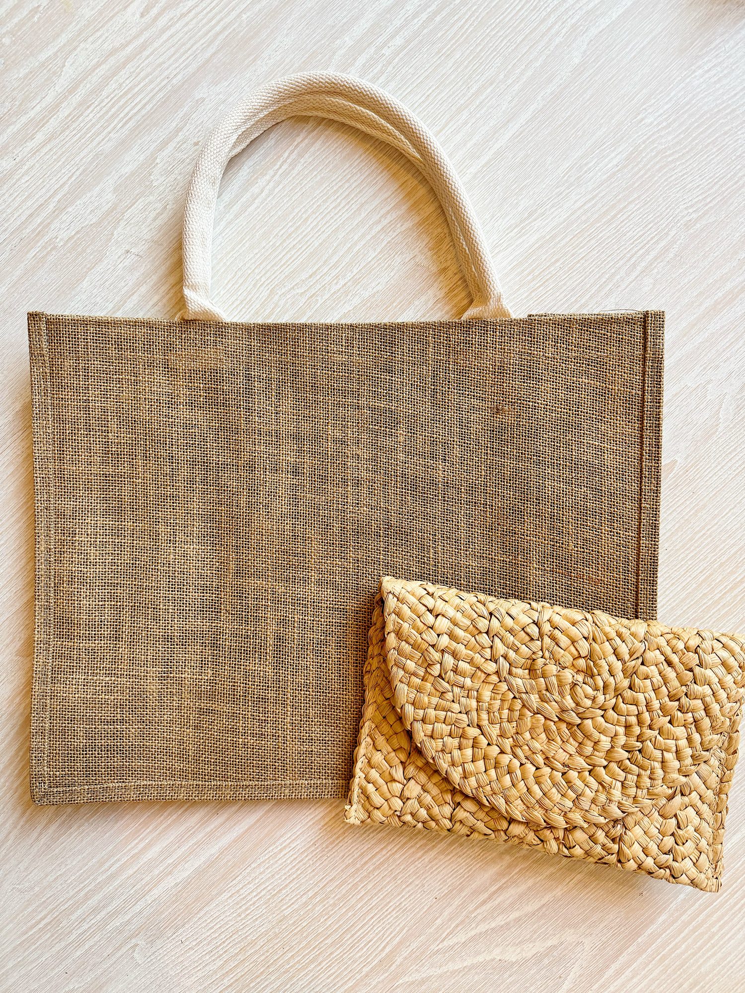 A rectangular jute tote bag with rope handles is placed next to a small woven straw clutch on a light wooden surface.