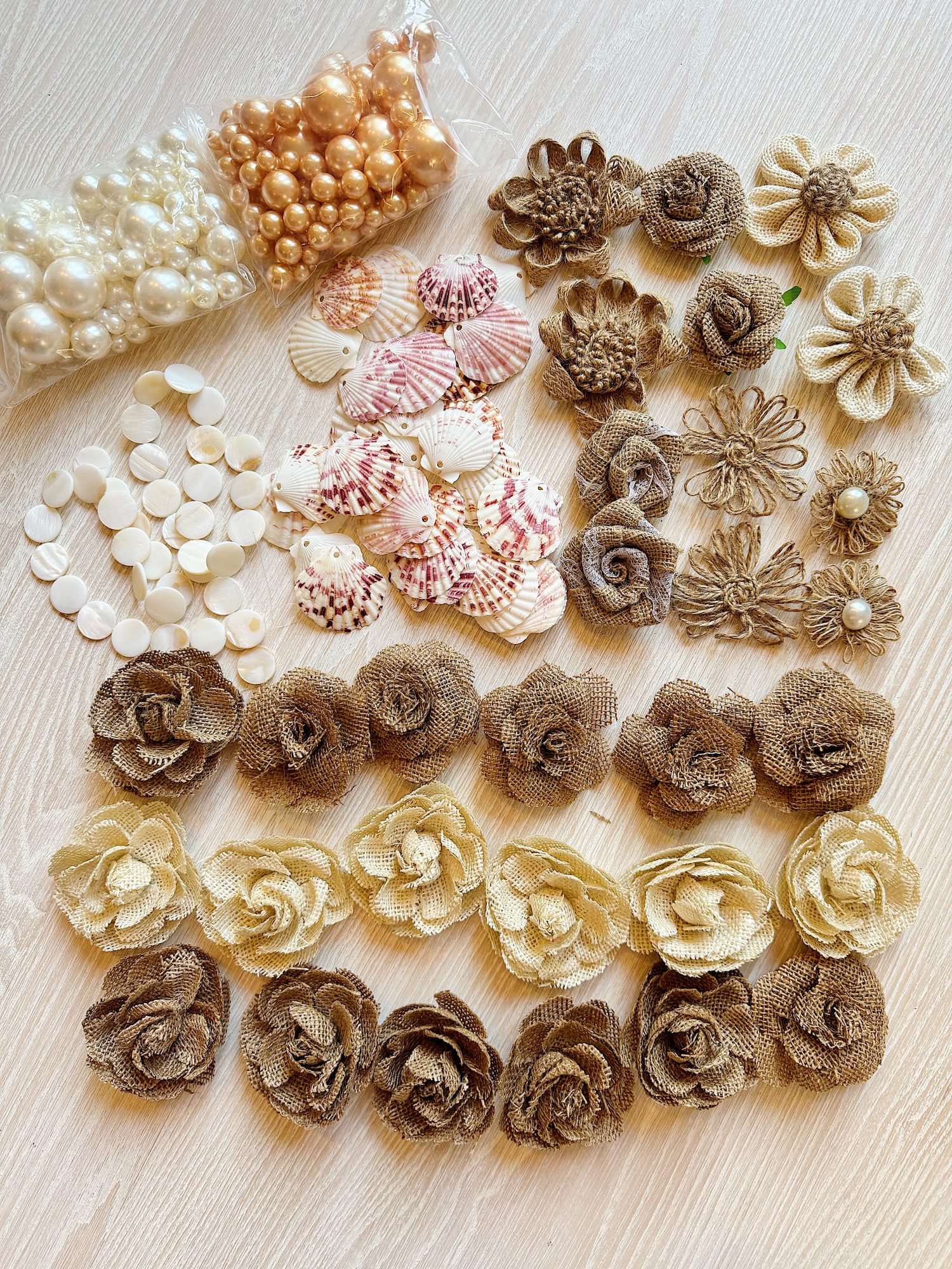Assorted craft materials on a light wooden surface, including faux pearls, seashells, white beads, and decorative fabric flowers in brown and beige tones arranged in groups.
