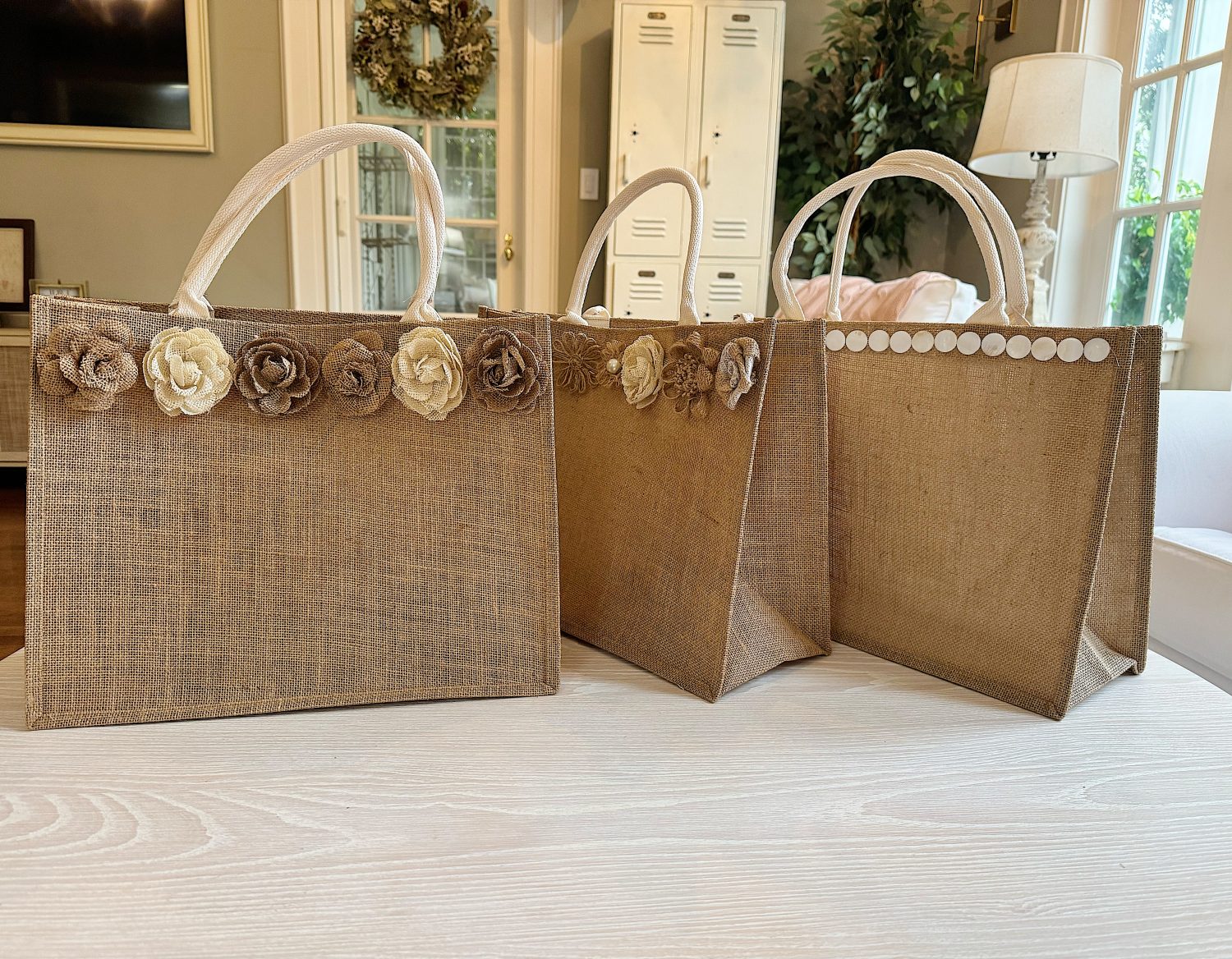 Three burlap tote bags are displayed on a light-colored table. Two bags are decorated with fabric flowers, and one is decorated with a row of white buttons.