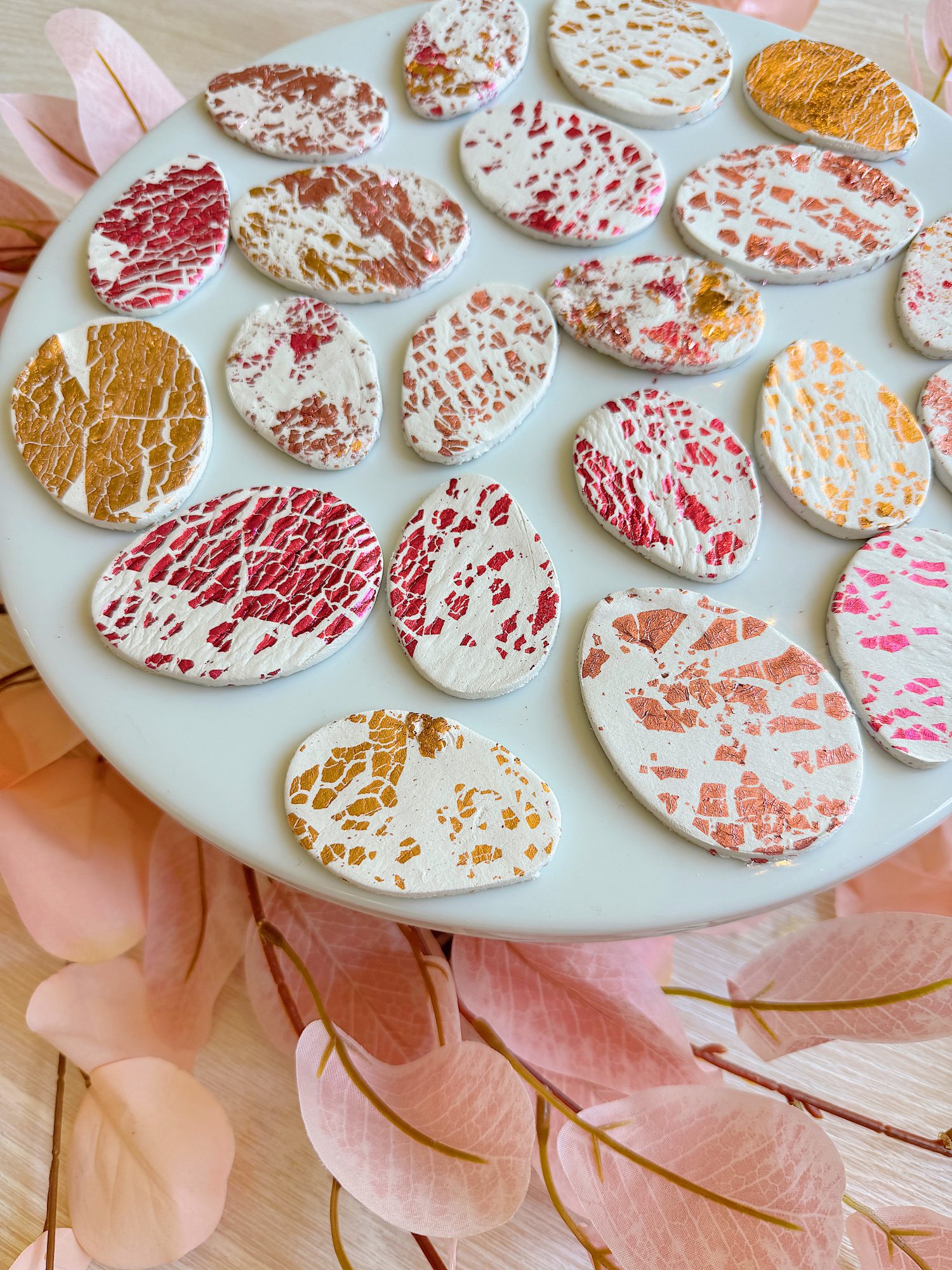 A white plate holds oval-shaped cookies with crackled patterns in red, pink, and gold. Pink translucent leaves are scattered around the plate.