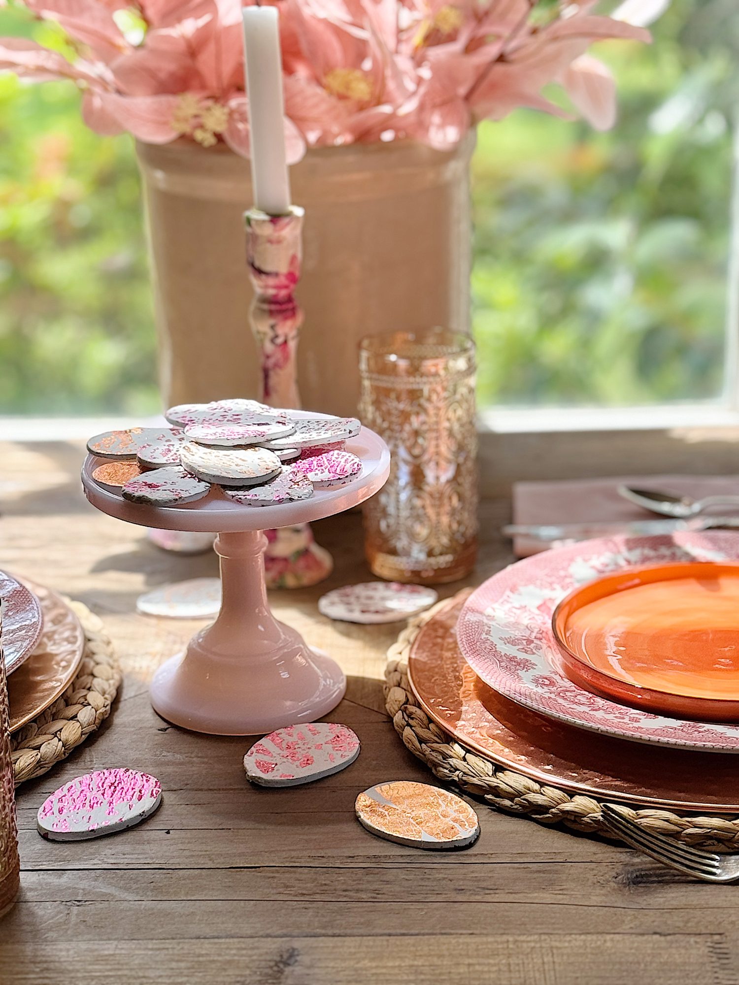 A table setup features pink flowers in a vase, decorative cookies on a stand, and orange and white floral plates with a wicker placemat. Candles and cutlery complete the arrangement.