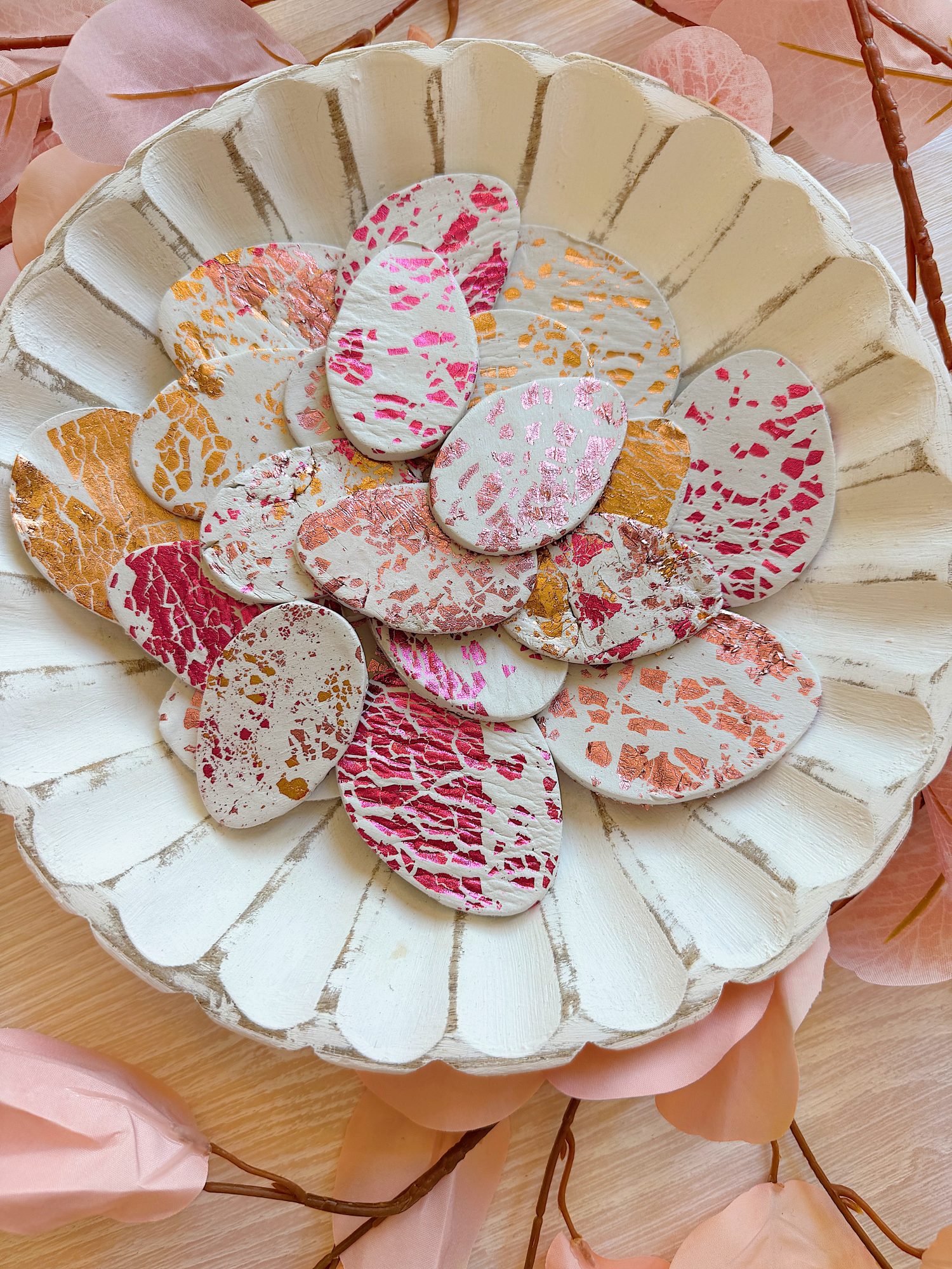 A white scalloped dish holds oval-shaped objects with pink, orange, and gold crackle patterns, surrounded by pink paper flowers.
