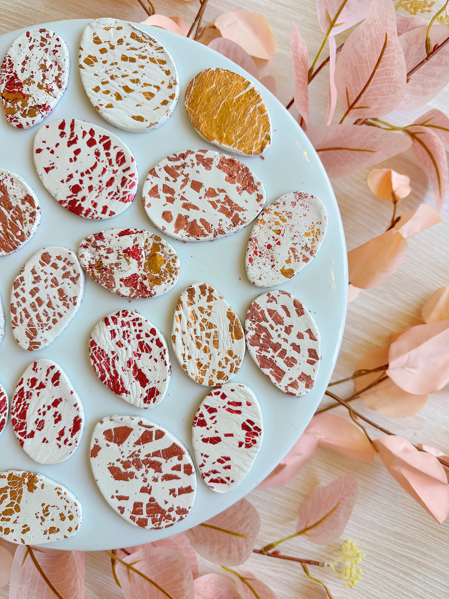 Oval-shaped cookies with white icing and red, pink, and gold splatter designs on a white plate. Surrounding the plate are scattered, pink decorative leaves on a light surface.