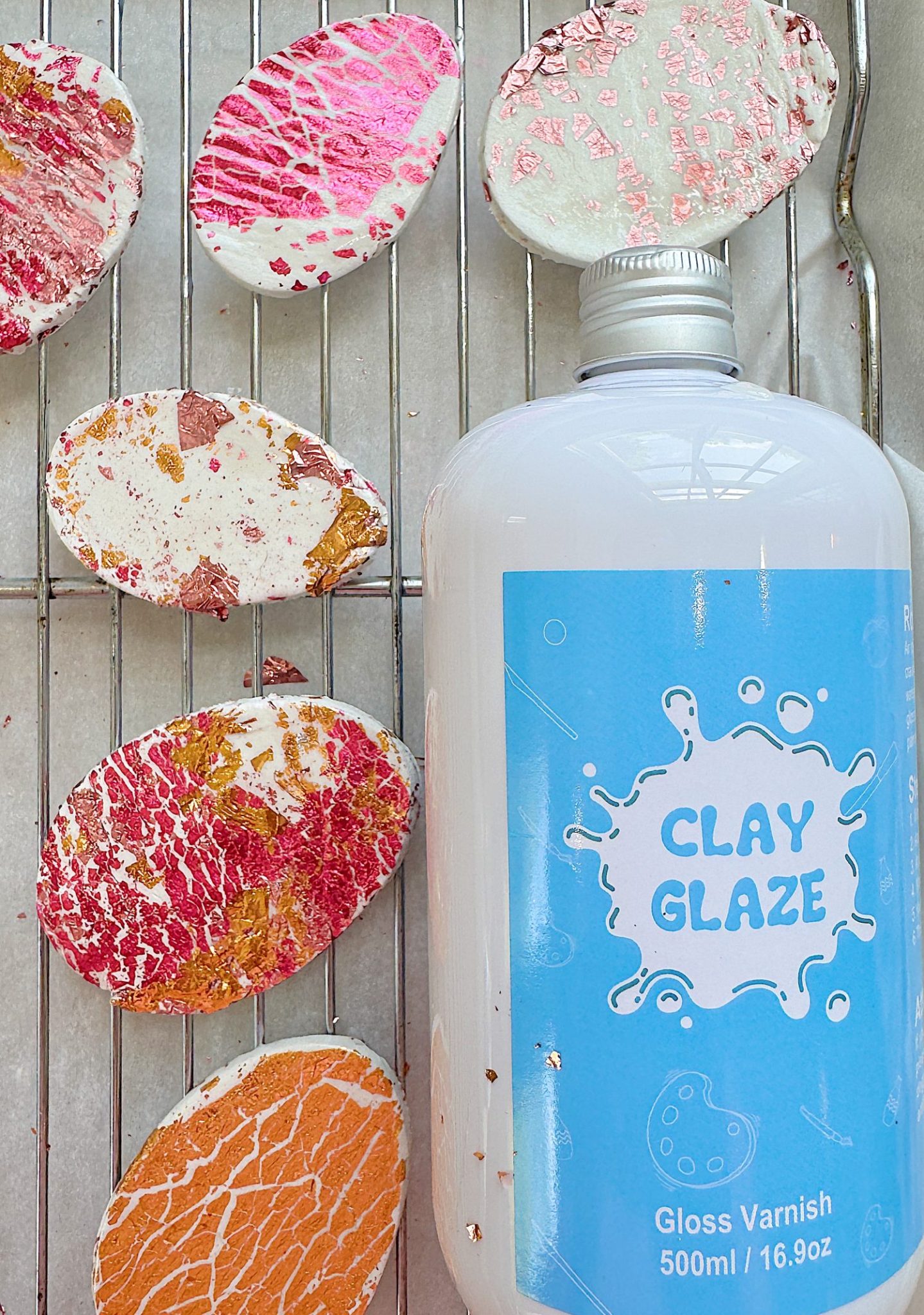 A blue bottle labeled "Clay Glaze" is next to several textured, multicolored clay pieces on a wire rack.