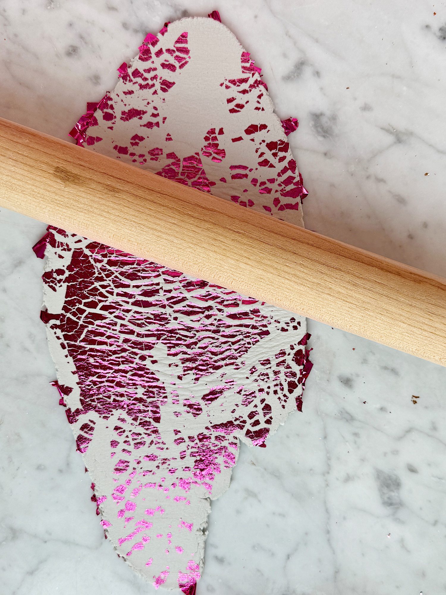A piece of pink and silver textured foil is being flattened by a wooden rolling pin on a marble surface.