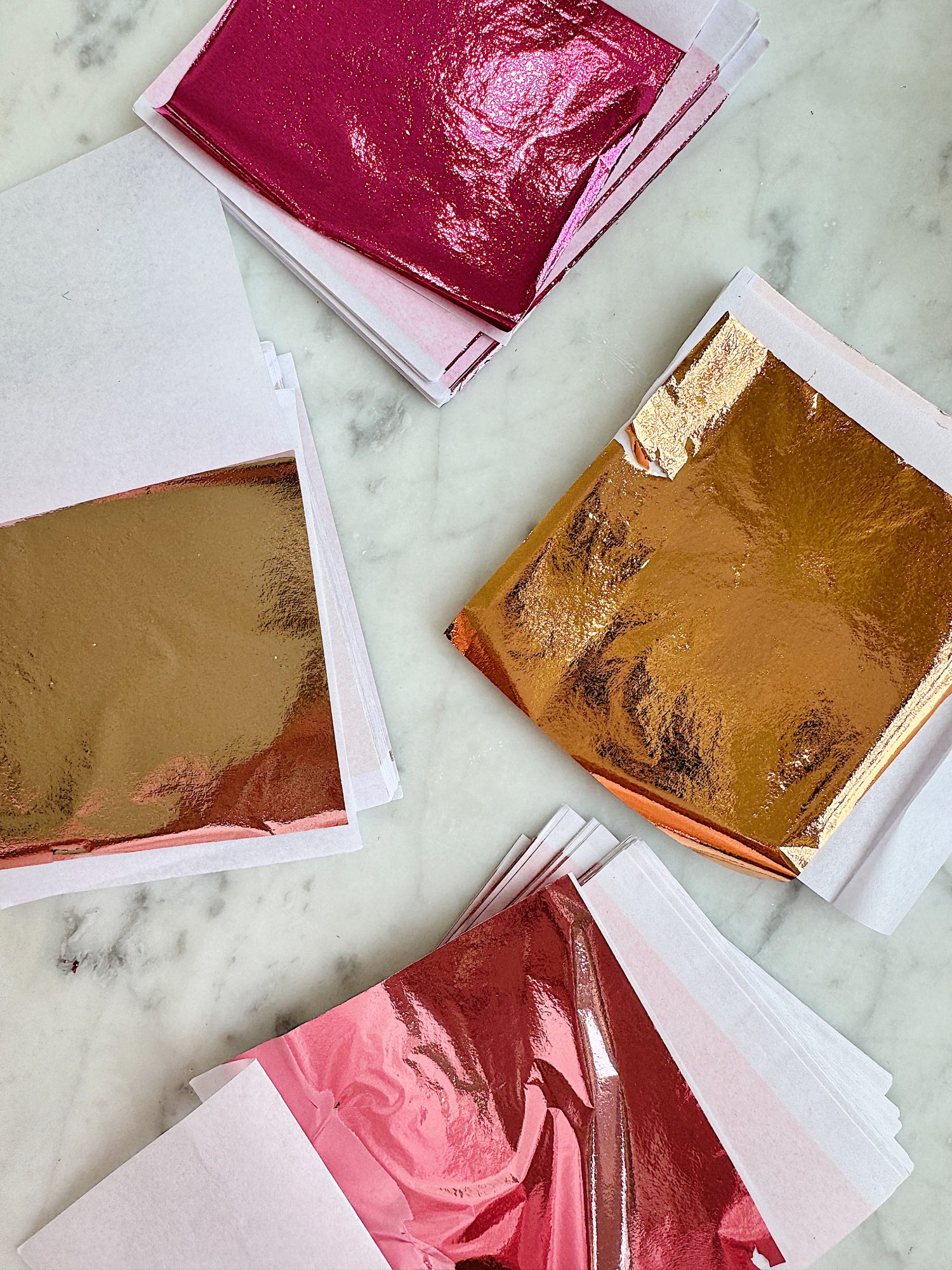 Stacks of sheets with metallic foil in pink, gold, and rose gold hues on a marble surface.
