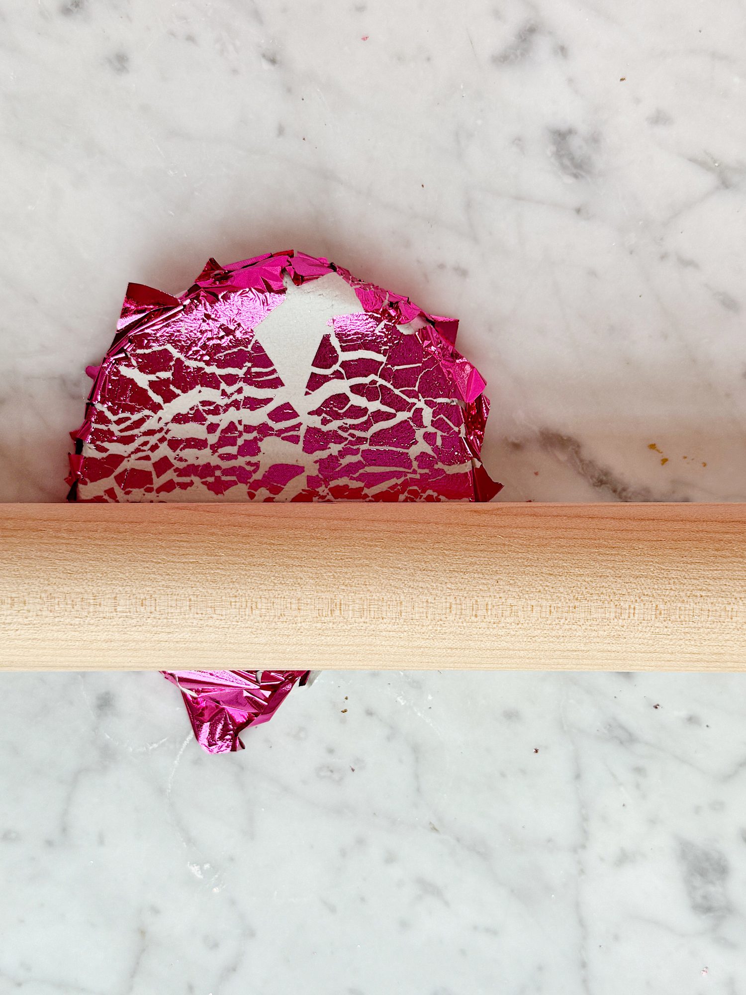 Rolling pin flattening pink foil wrap on marble surface.