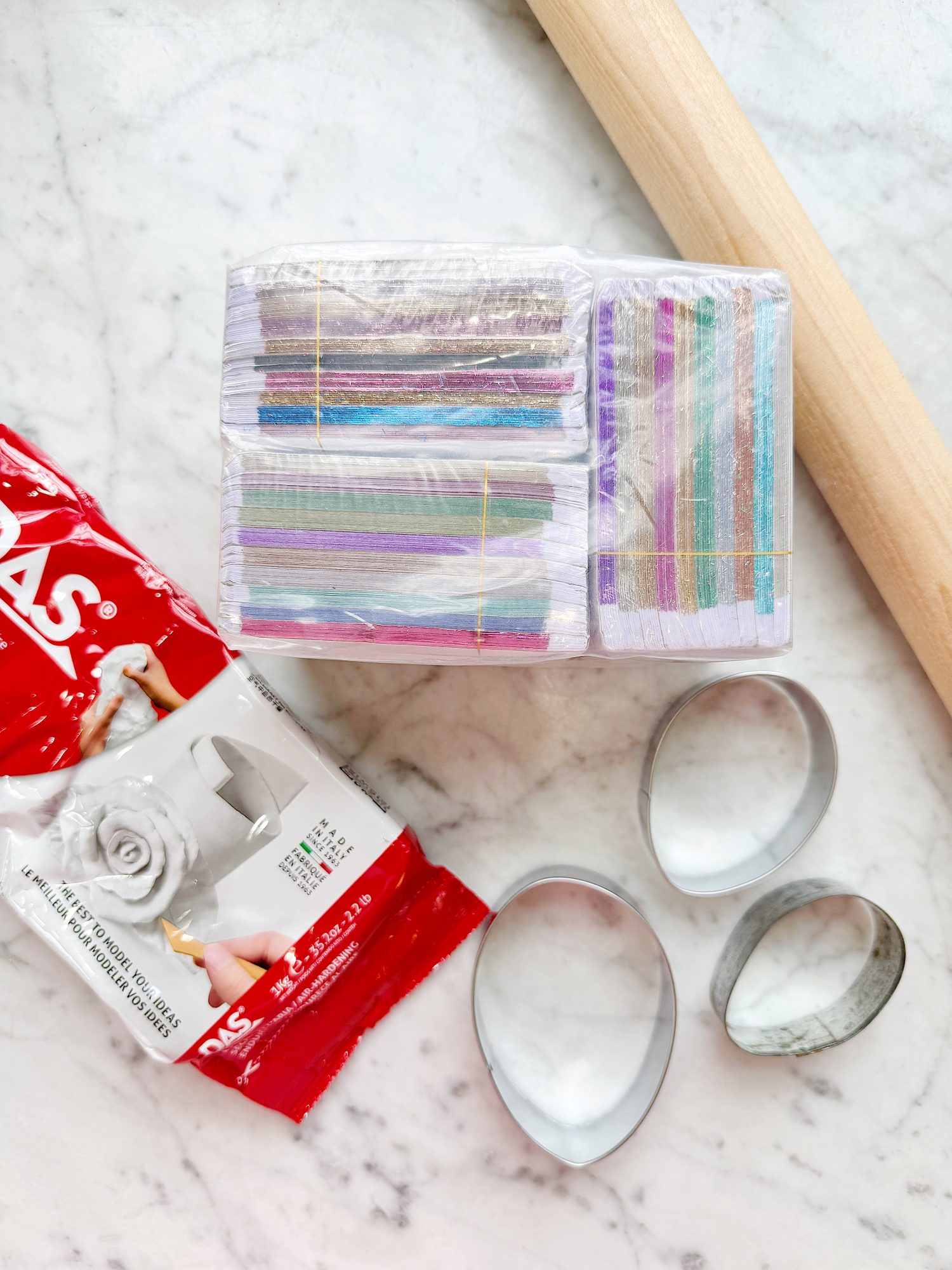 Crafting supplies on a marble surface, including a wooden rolling pin, colorful stacked sheets, aluminum cutters, and a red pack of white soft modeling clay.