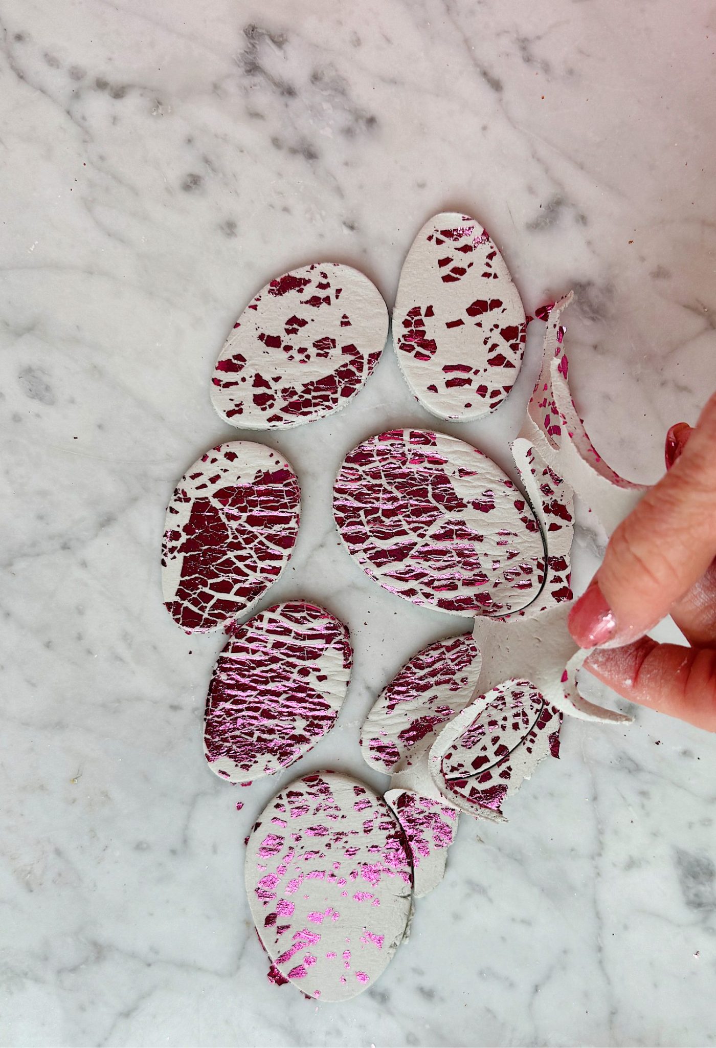 Hand holding a piece of cracked eggshell painted white with pink cracks on a marble surface. Several painted eggshell pieces are scattered around.