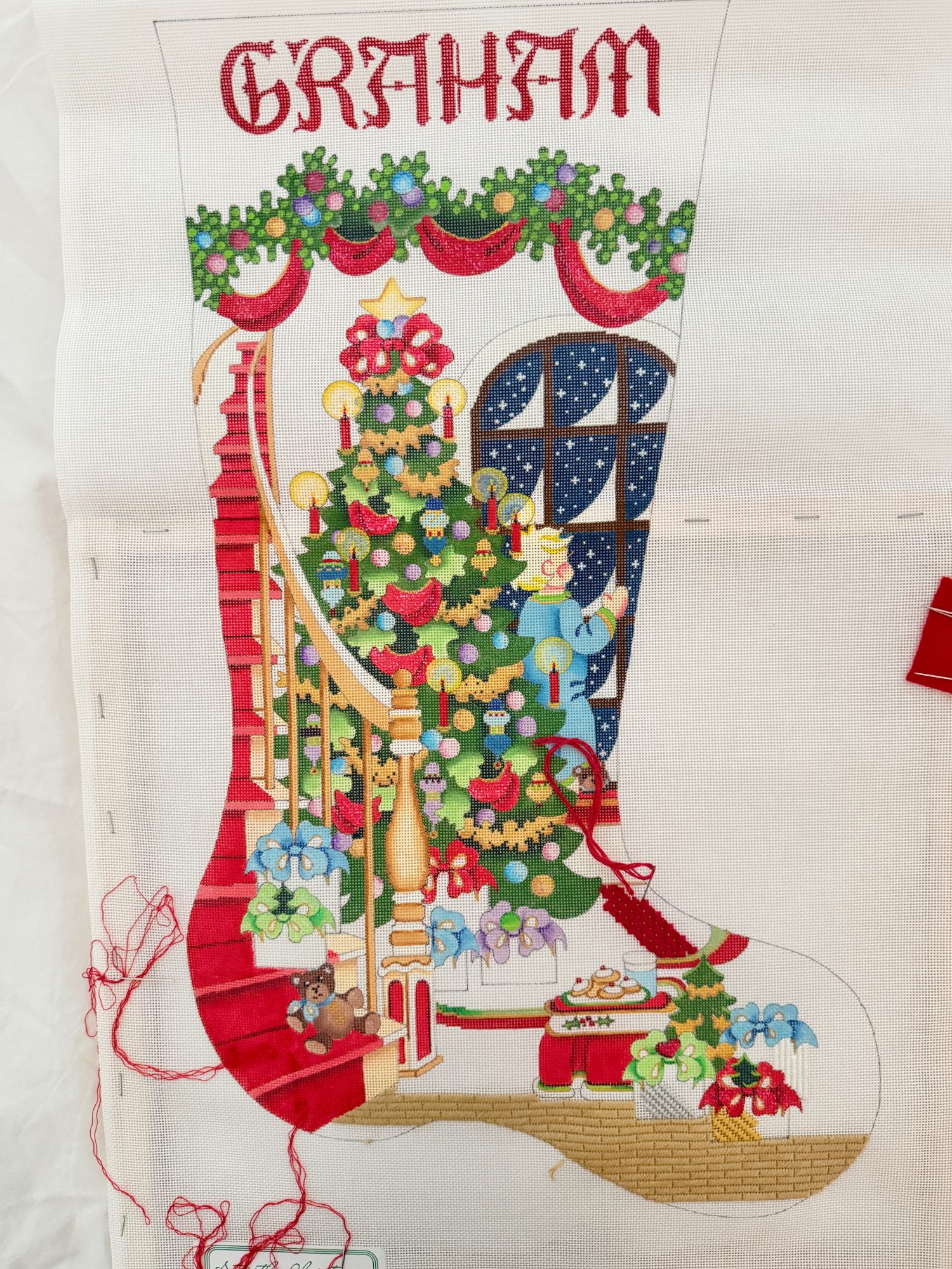 A Christmas stocking cross-stitch featuring a decorated tree, staircase, gifts, and a teddy bear, with red trim and the name "GRAHAM" embroidered at the top.