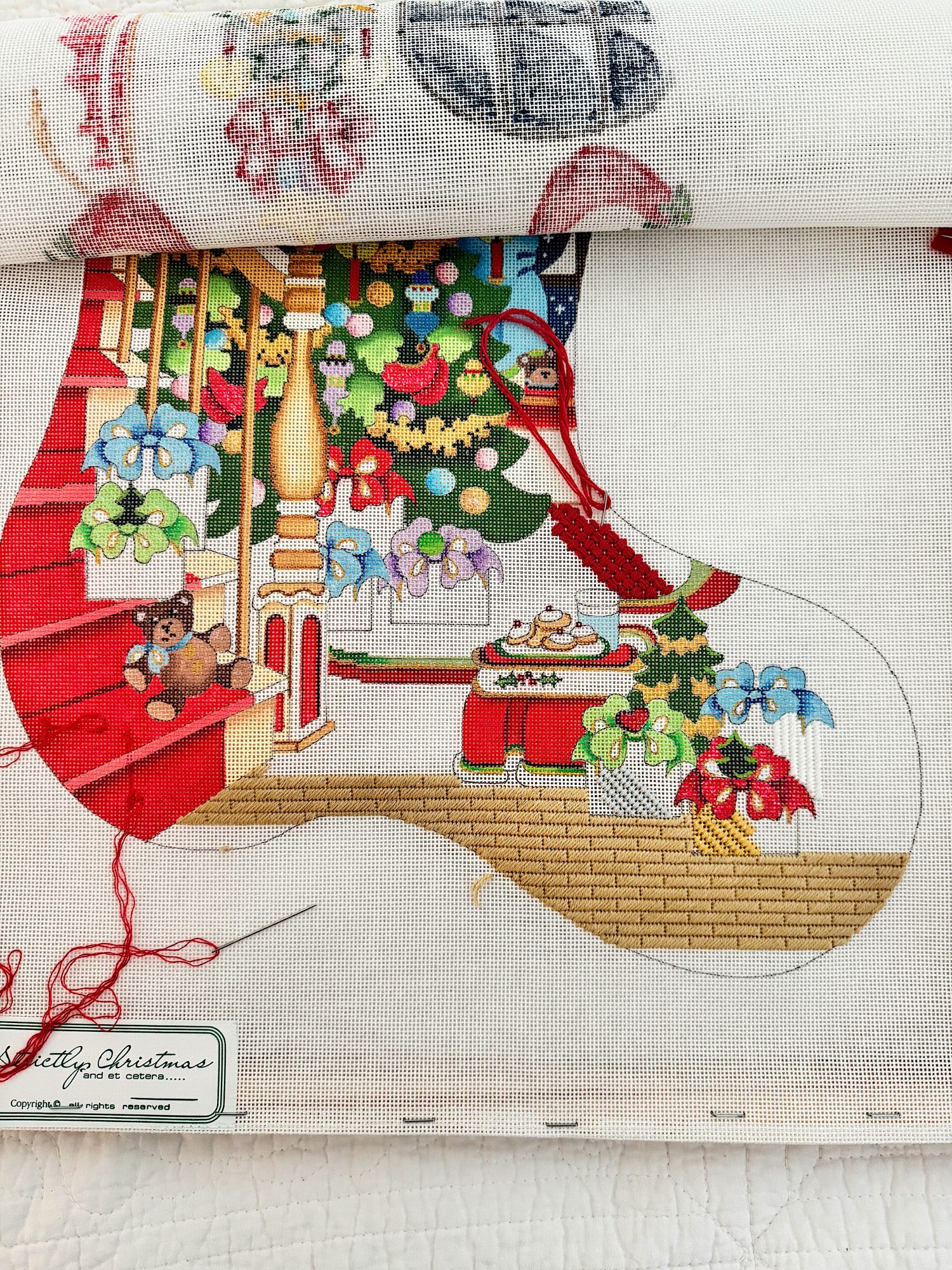 Needlepoint canvas depicting a Christmas stocking design with gifts, a teddy bear, and holiday decorations in progress.