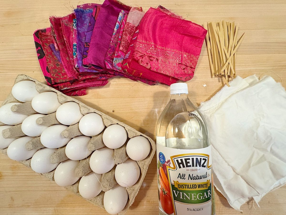 A carton of eggs, fabric pieces, a bottle of white vinegar, wooden skewers, and white cloth are arranged on a wooden surface.