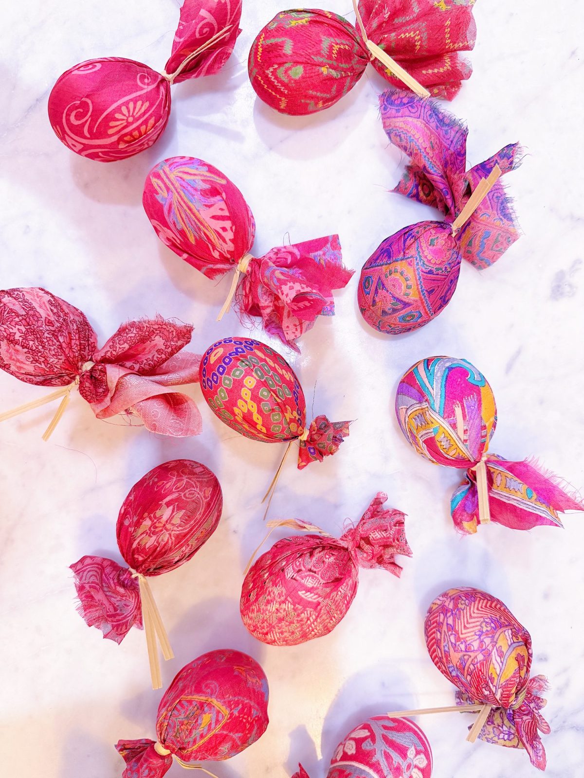 Assorted vibrant eggs wrapped in patterned pink and red cloth with tied ends, placed on a white marble surface.