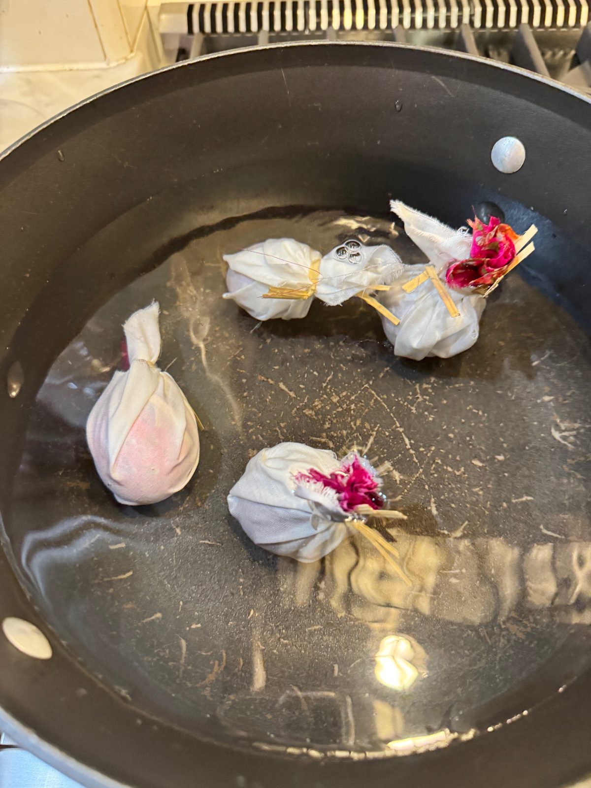 Four fabric bundles tied with rubber bands are simmering in a pot of water on a stove.