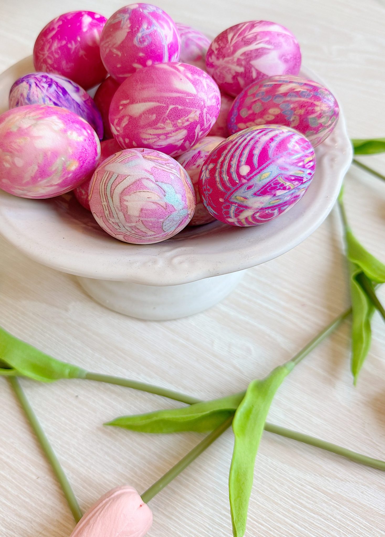 A white bowl filled with pink and white marbled eggs rests on a light wood surface. Green leaves and a pink flower are placed nearby.