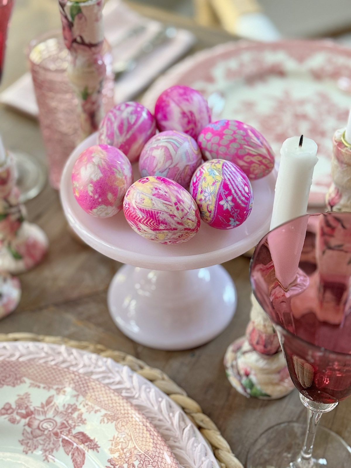 A table setting with a pedestal holding pink patterned eggs, surrounded by floral plates, a candle, and a pink glass.