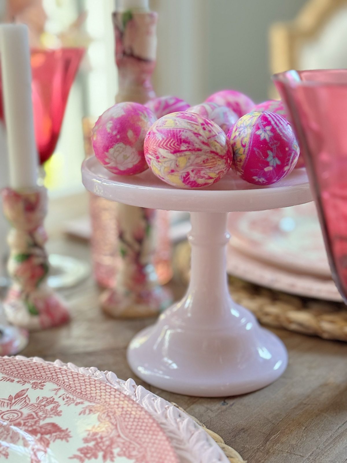A white cake stand holds decorative pink and floral-patterned eggs. Nearby, pink patterned glassware and plates are arranged on a wooden table.