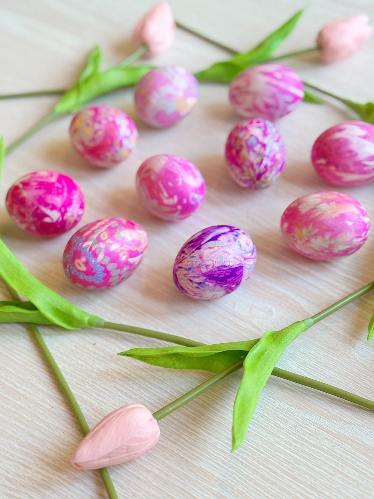 Pink and purple marbled eggs arranged in a square with pink tulip buds and green leaves at each corner on a light wooden surface.