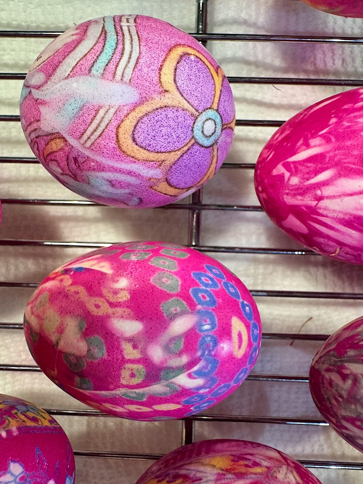 Colorful, intricately patterned Easter eggs drying on a metal rack, with floral and abstract designs in pink, purple, and blue tones.