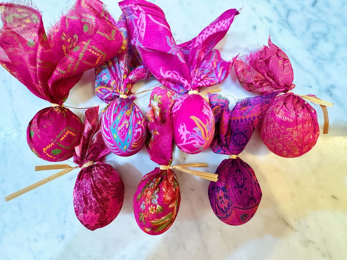 Ten decorative eggs wrapped in vibrant pink and purple fabric with patterns, tied with straw, arranged on a marble surface.