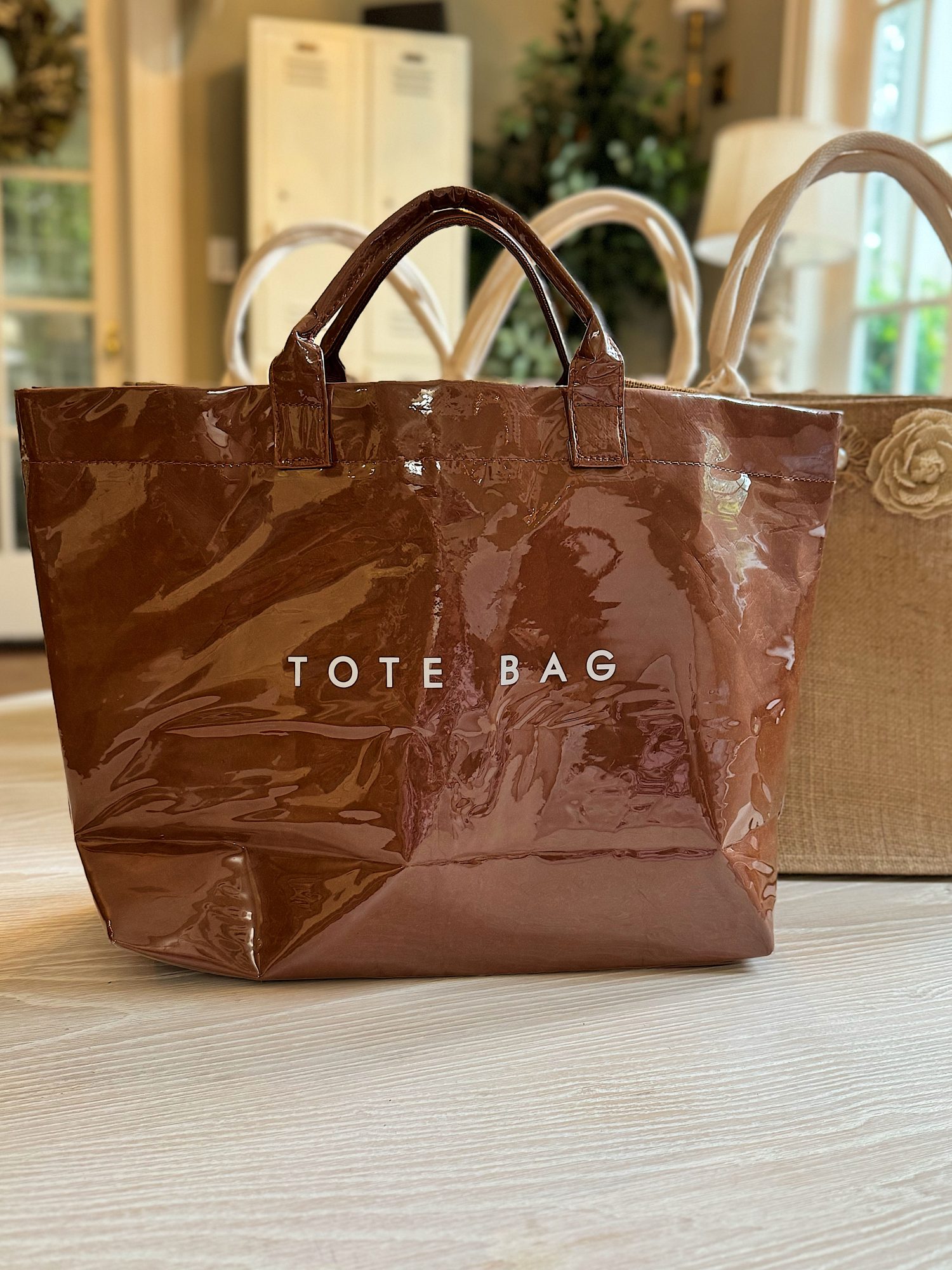 A brown, glossy tote bag labeled "TOTE BAG" sits on a light-colored surface next to a tan woven bag with a flower detail.