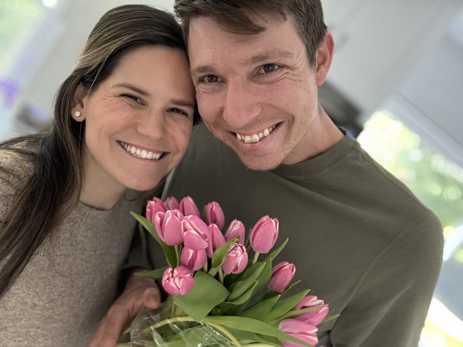 A smiling couple poses closely together indoors, holding a bouquet of pink tulips.
