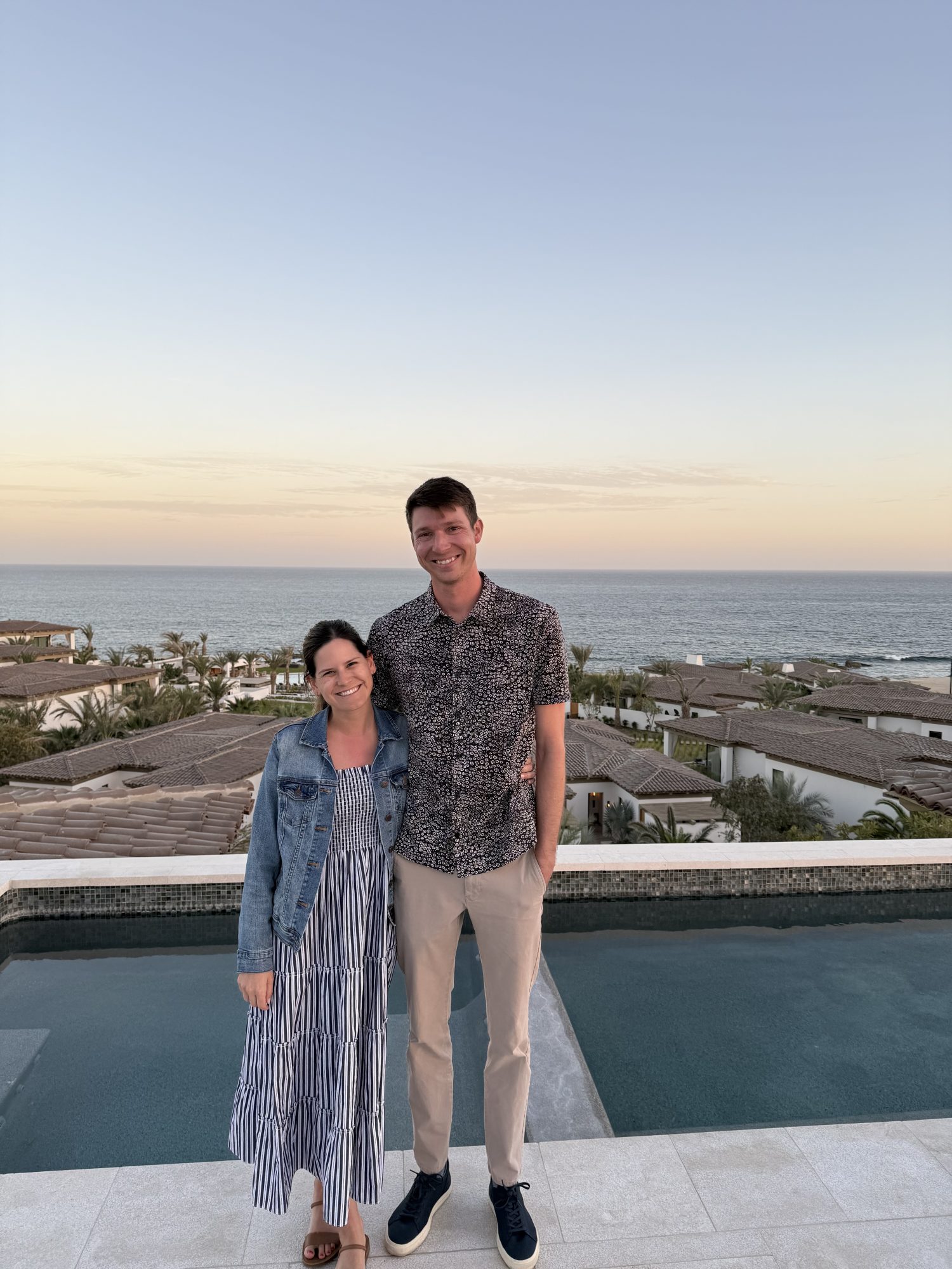 A woman and man stand side by side, smiling in front of a pool with a view of rooftops, palm trees, and the ocean at sunset.