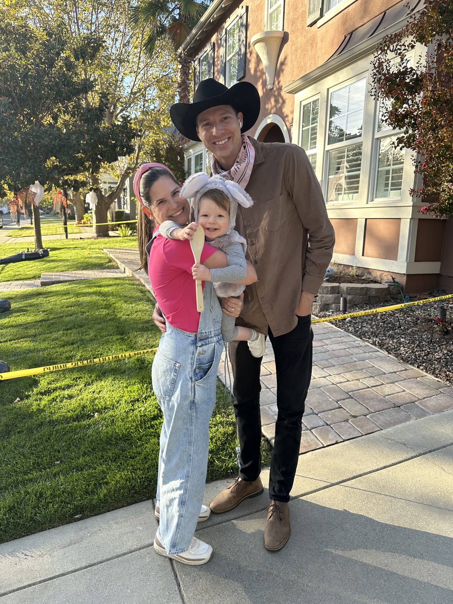 A man in a black cowboy hat stands next to a woman in a pink top and jeans holding a smiling child wearing bunny ears on a sunny sidewalk in front of a house.