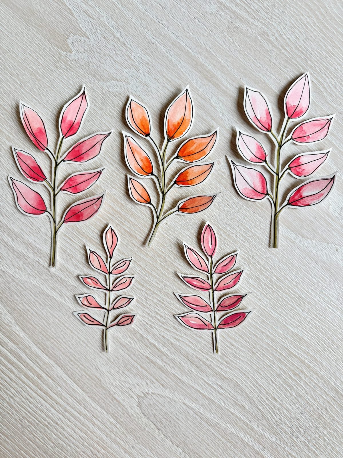 Five hand-drawn and watercolor-painted paper cutouts of leafy branches in shades of red, pink, and orange are arranged on a light wood surface.