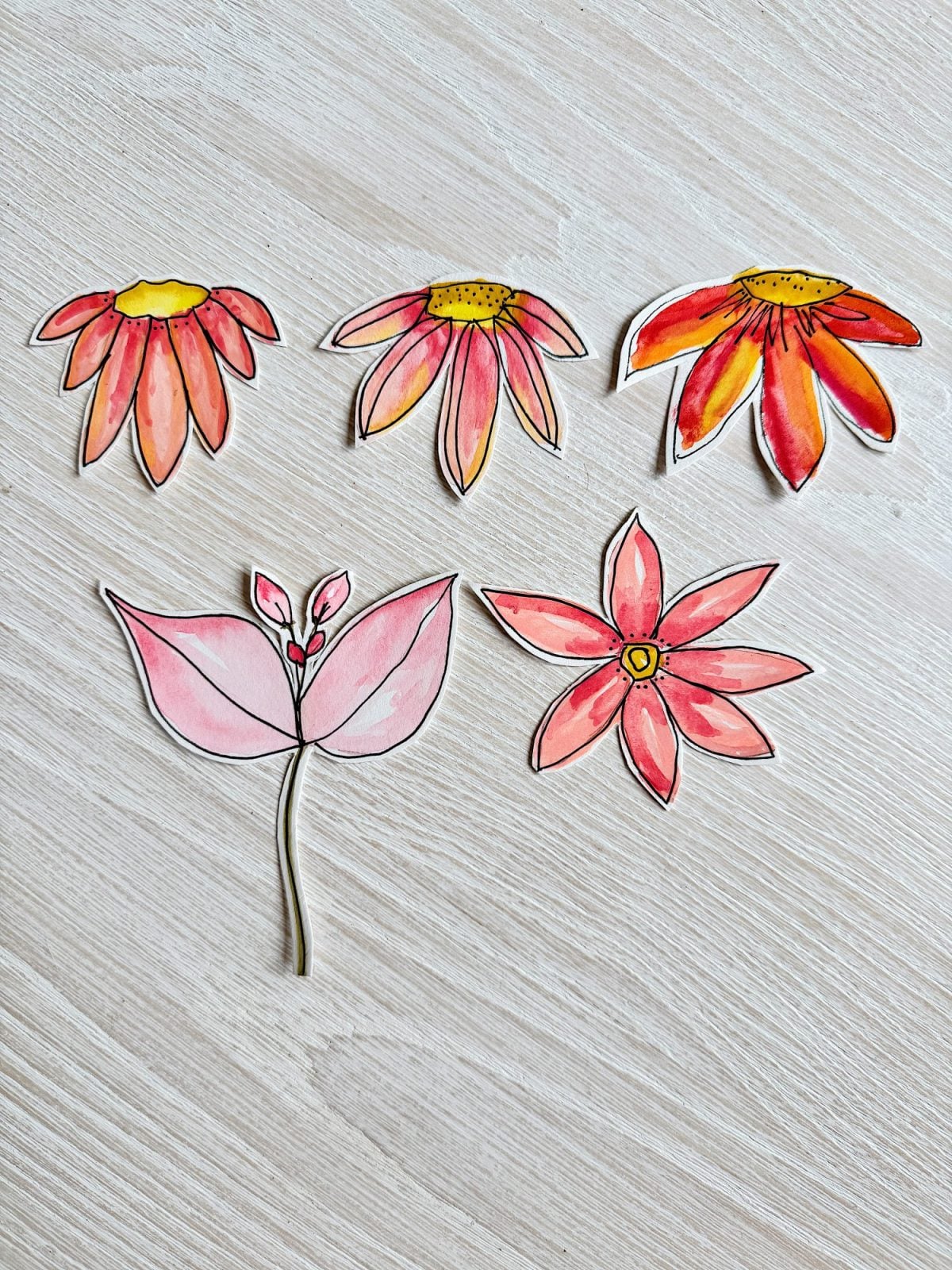 Five hand-drawn and painted cutout flowers with pink and yellow tones are arranged on a light wooden surface.