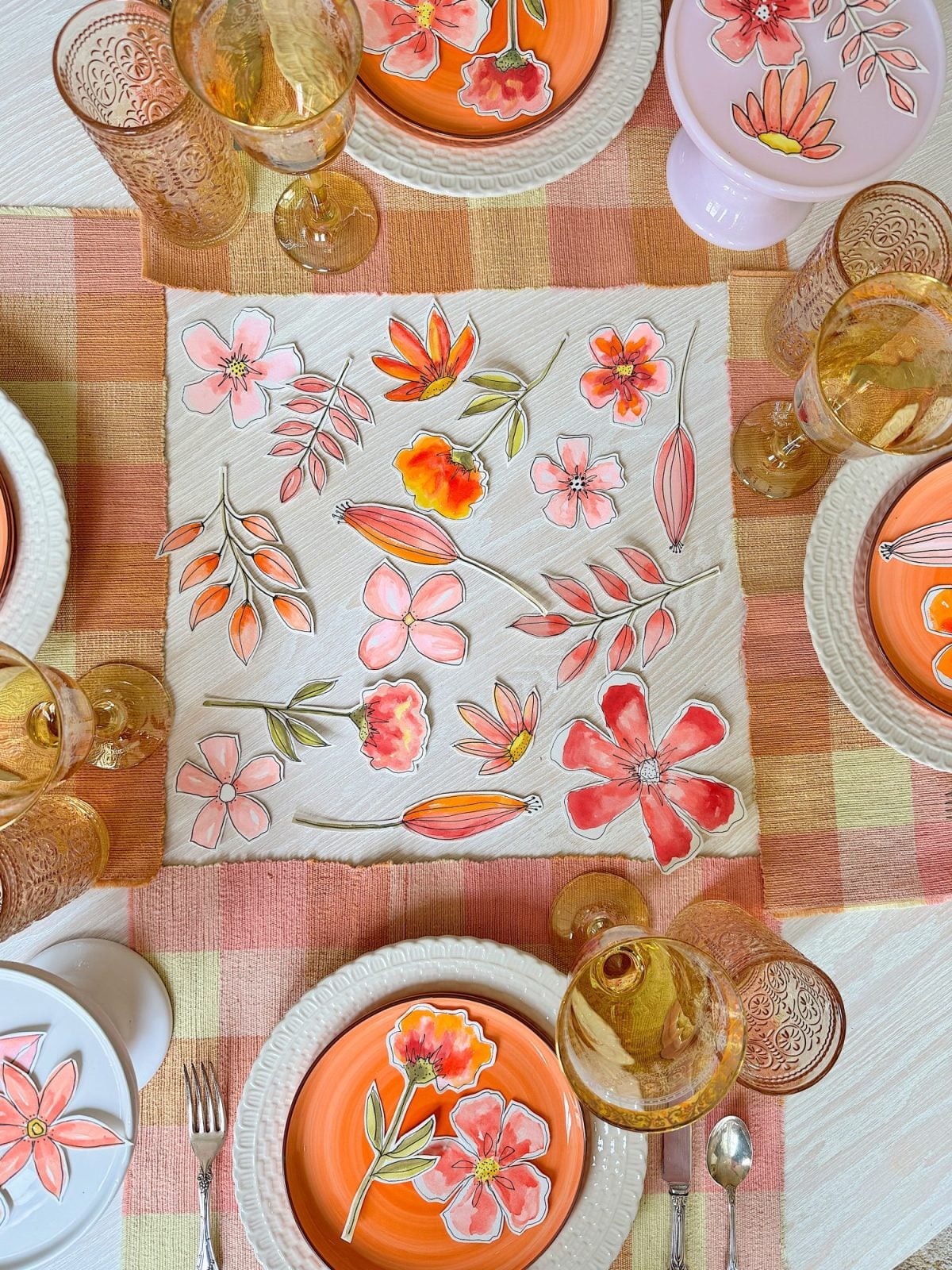 A table set with orange and pink floral plates, matching glassware, silverware, and a floral-patterned napkin on a checked tablecloth.