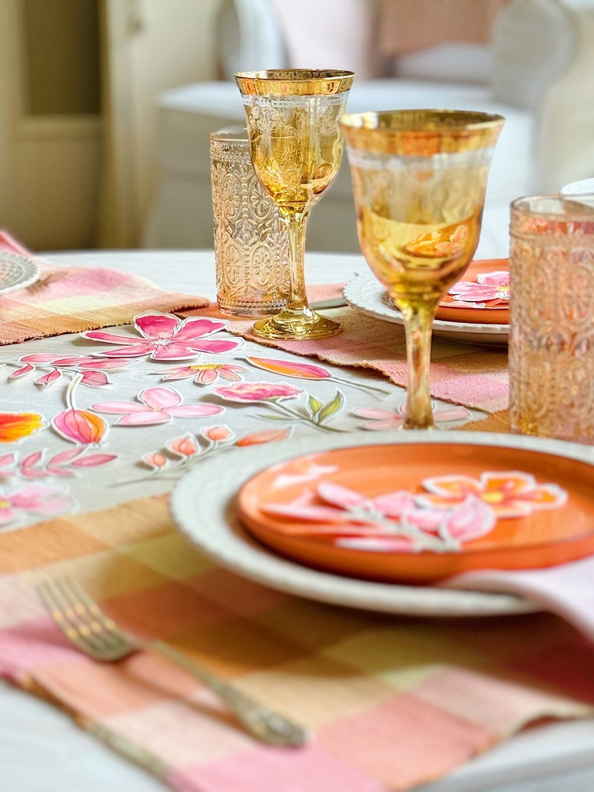 A close-up of a table setting with floral plates, gold-rimmed glasses, patterned tumblers, and pink and orange placemats.