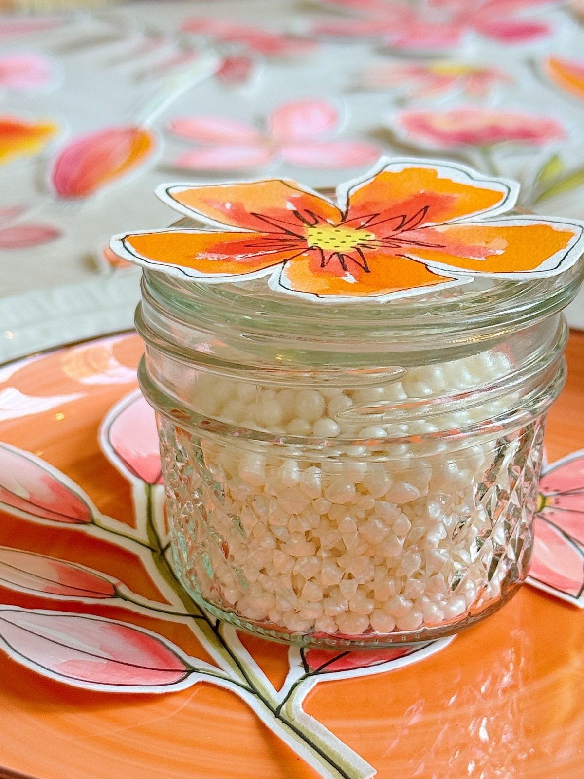 A small glass jar filled with white beads sits on an orange plate with a colorful floral paper decoration on top.