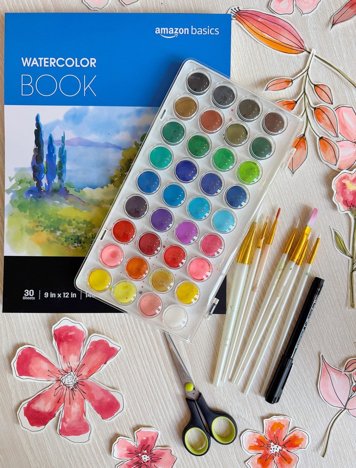 A watercolor paint set, four paintbrushes, scissors, and paper flower cutouts are arranged on top of a watercolor book with a painted landscape on the cover.