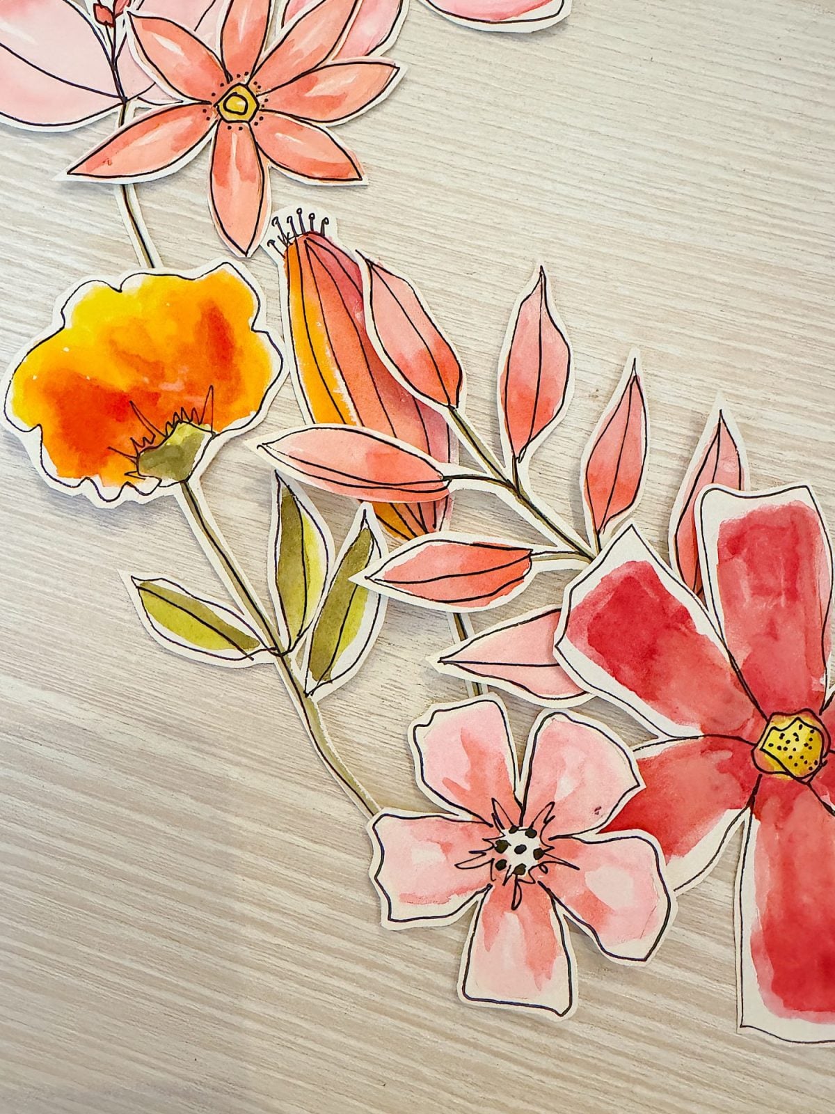 Watercolor illustrations of various pink, orange, and yellow flowers with leaves are cut out and arranged on a light wooden surface.