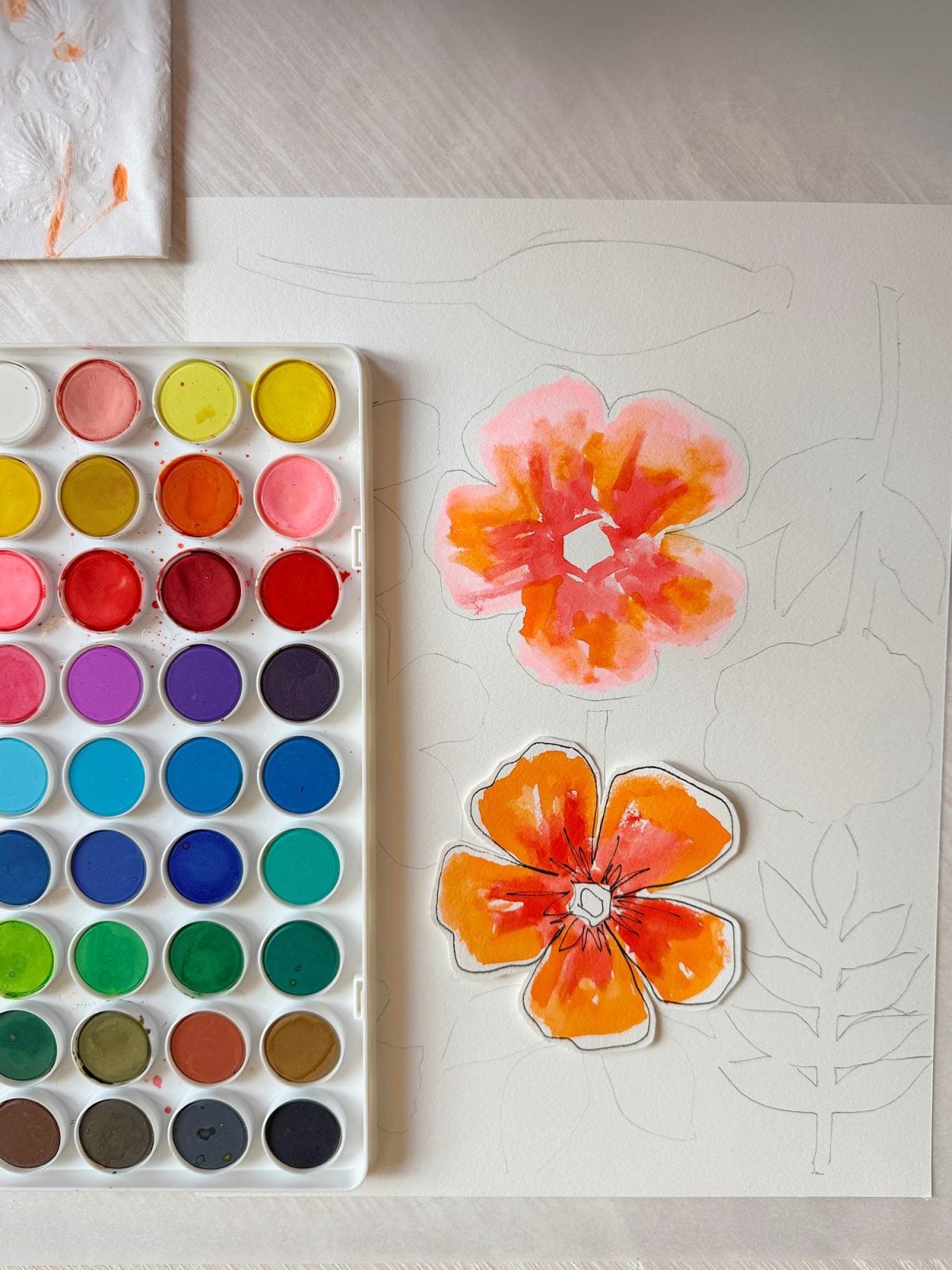 An open watercolor paint palette sits next to a sheet of paper with two colorful painted flowers and some outlined floral sketches.