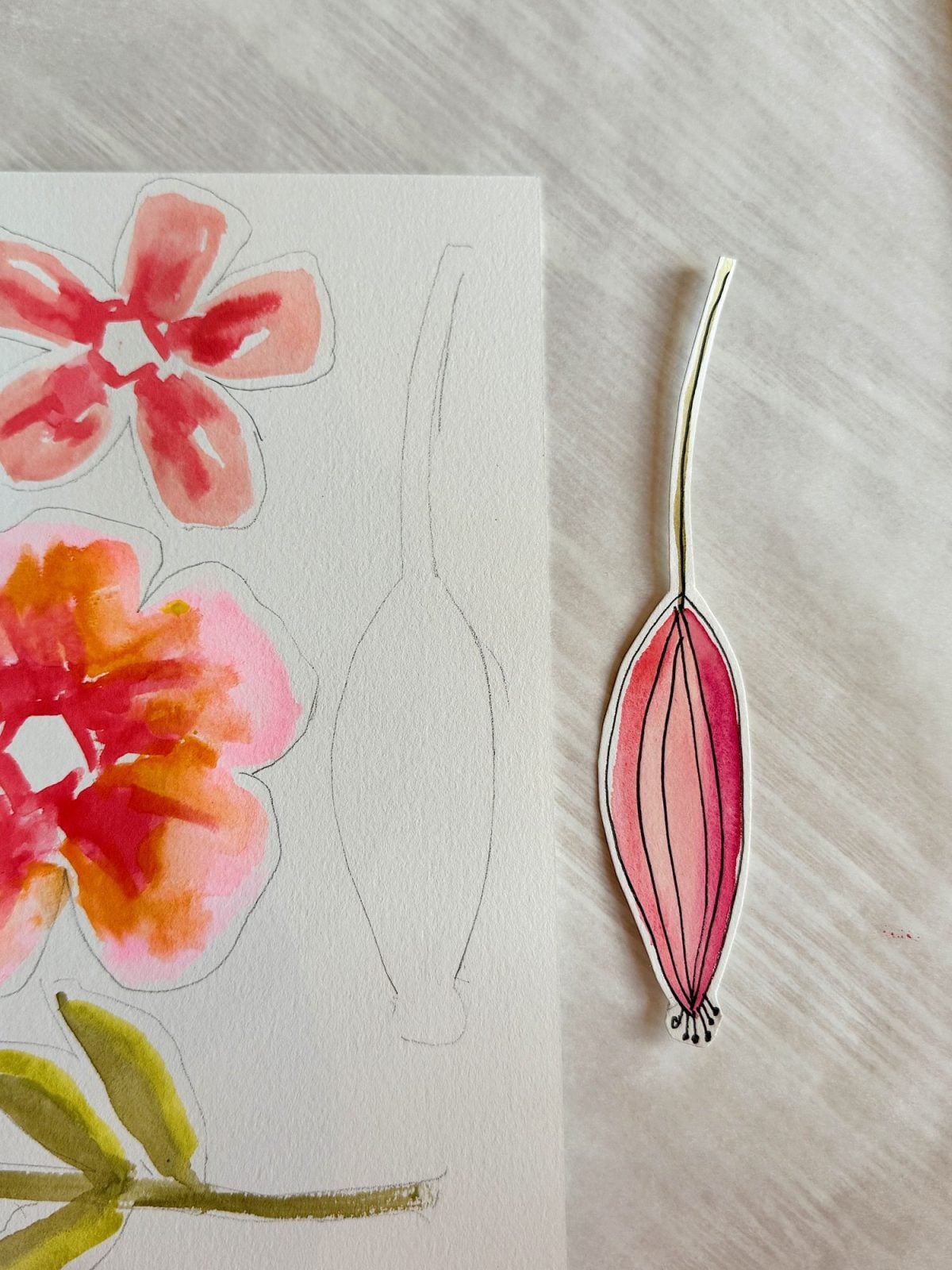 A piece of paper with watercolor flower sketches and an outlined bud; a separate cut-out drawing of a pink flower bud is placed to the right on a light surface.