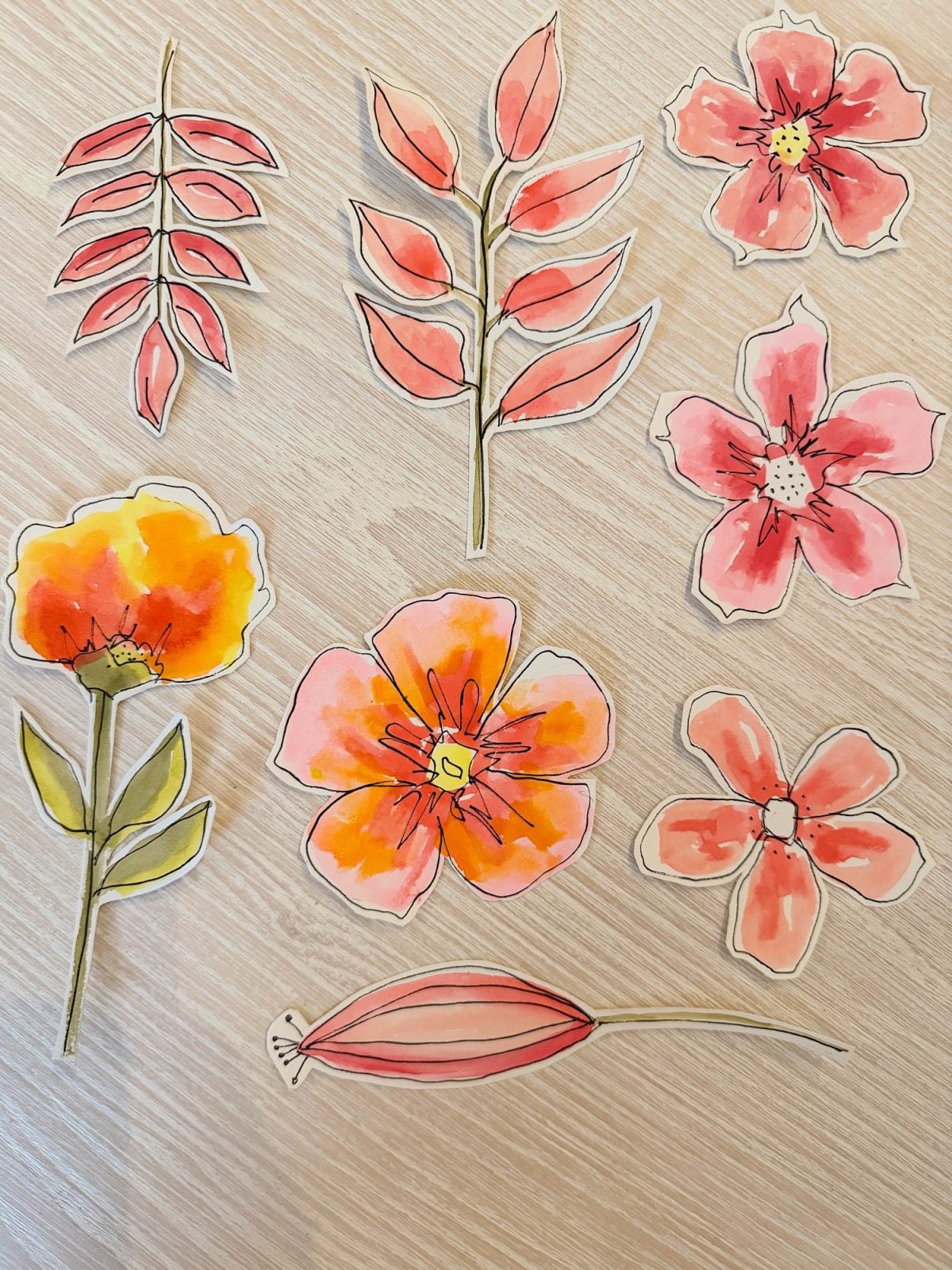 Hand-drawn and watercolor-painted cutouts of various stylized flowers and leaves are arranged on a light wooden surface.