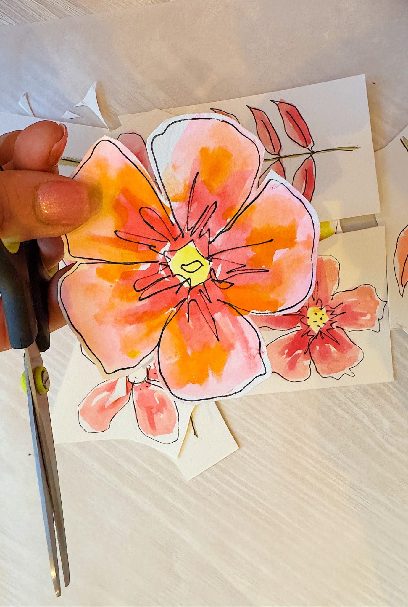 A hand holding scissors cuts out a drawing of orange and pink flowers on white paper placed on a light surface.